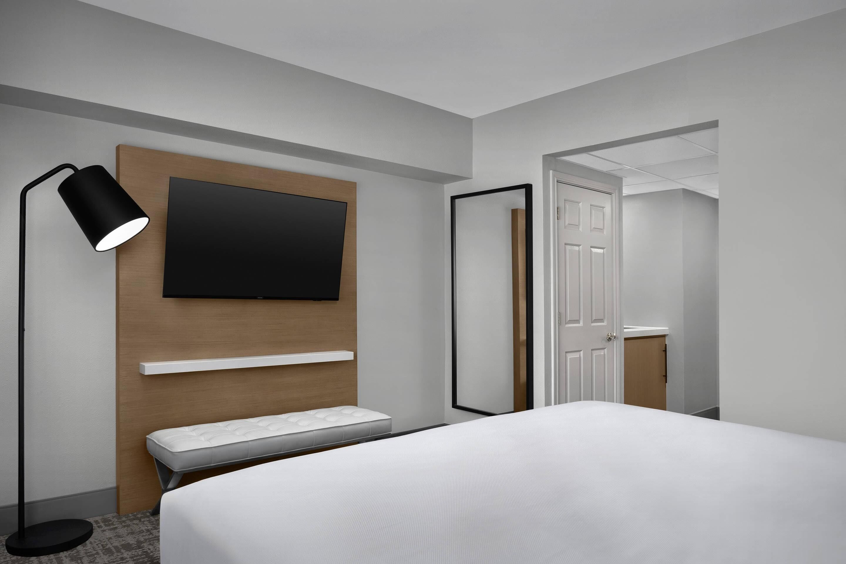 A modern hotel room with a bed, a bedside table, a flat-screen TV mounted on a wooden panel, a floor lamp, and a mirror.