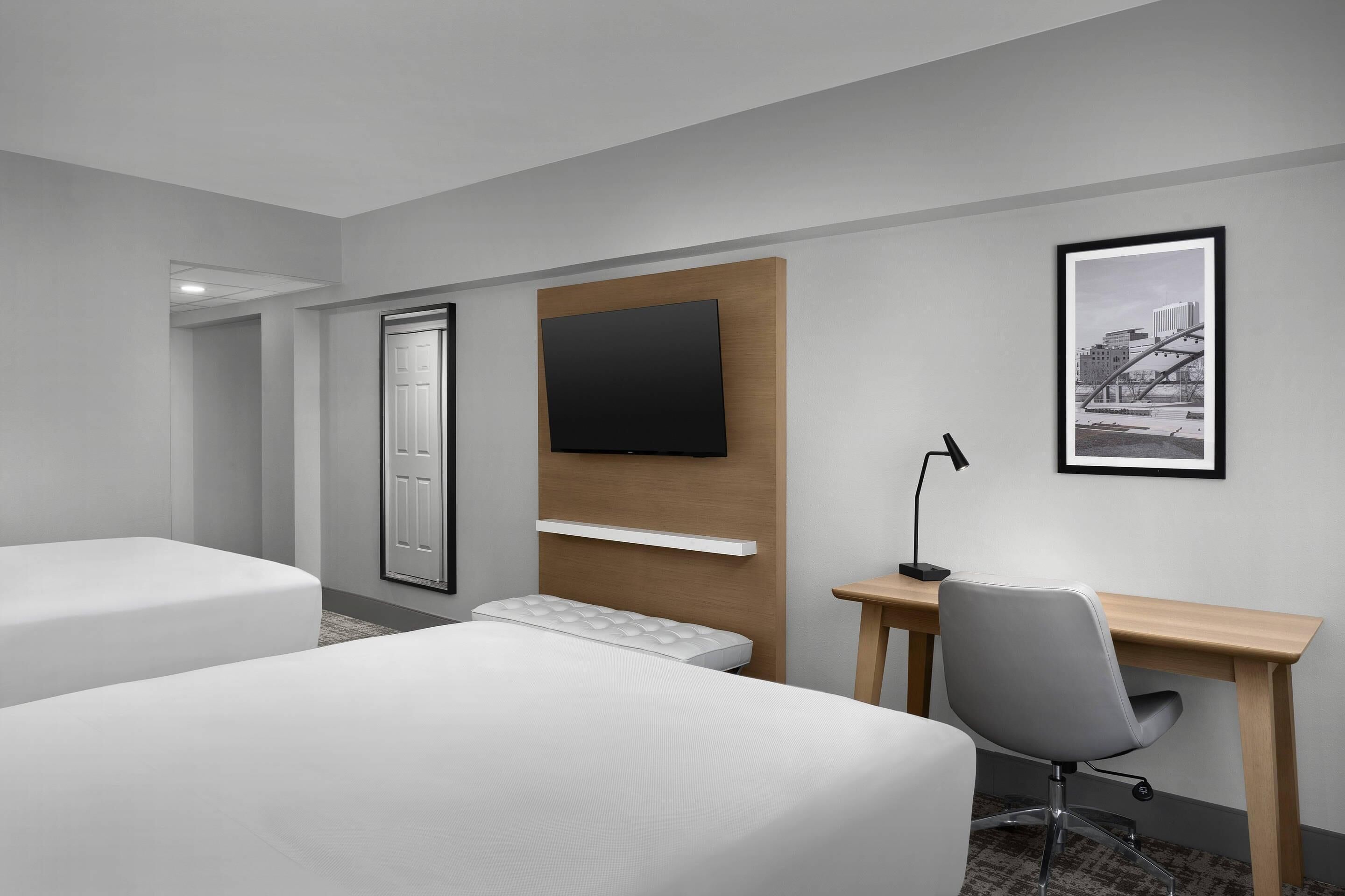 A hotel room with two beds, a desk, a chair, a TV mounted on the wall, and a framed picture.