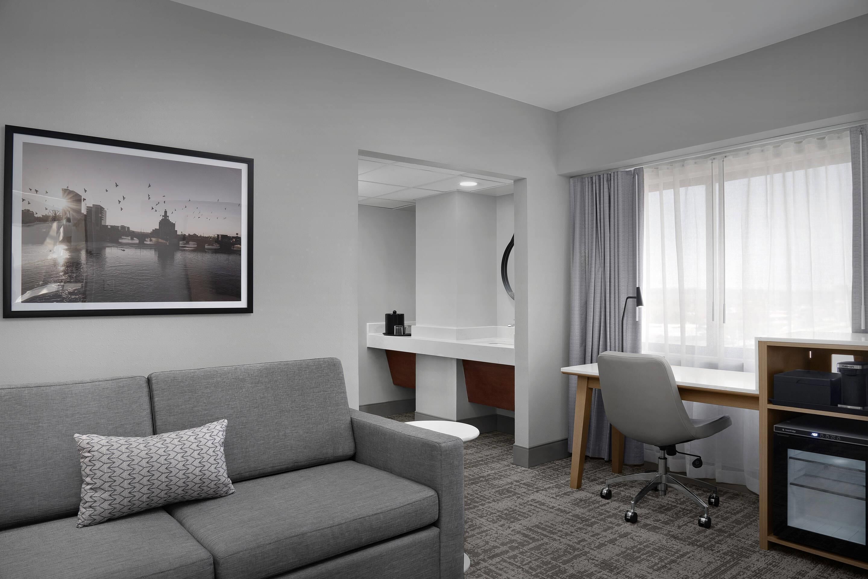 Ein Hotelzimmer mit grauem Sofa, Schreibtisch, Stuhl, Drucker und einem gerahmten Bild an der Wand.