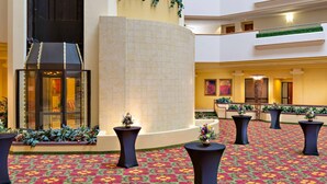 A hotel lobby with a patterned carpet, black tables with floral centerpieces, a glass elevator, and a tiled column. - Radisson Hotel Cedar Rapids (Cedar Rapids)