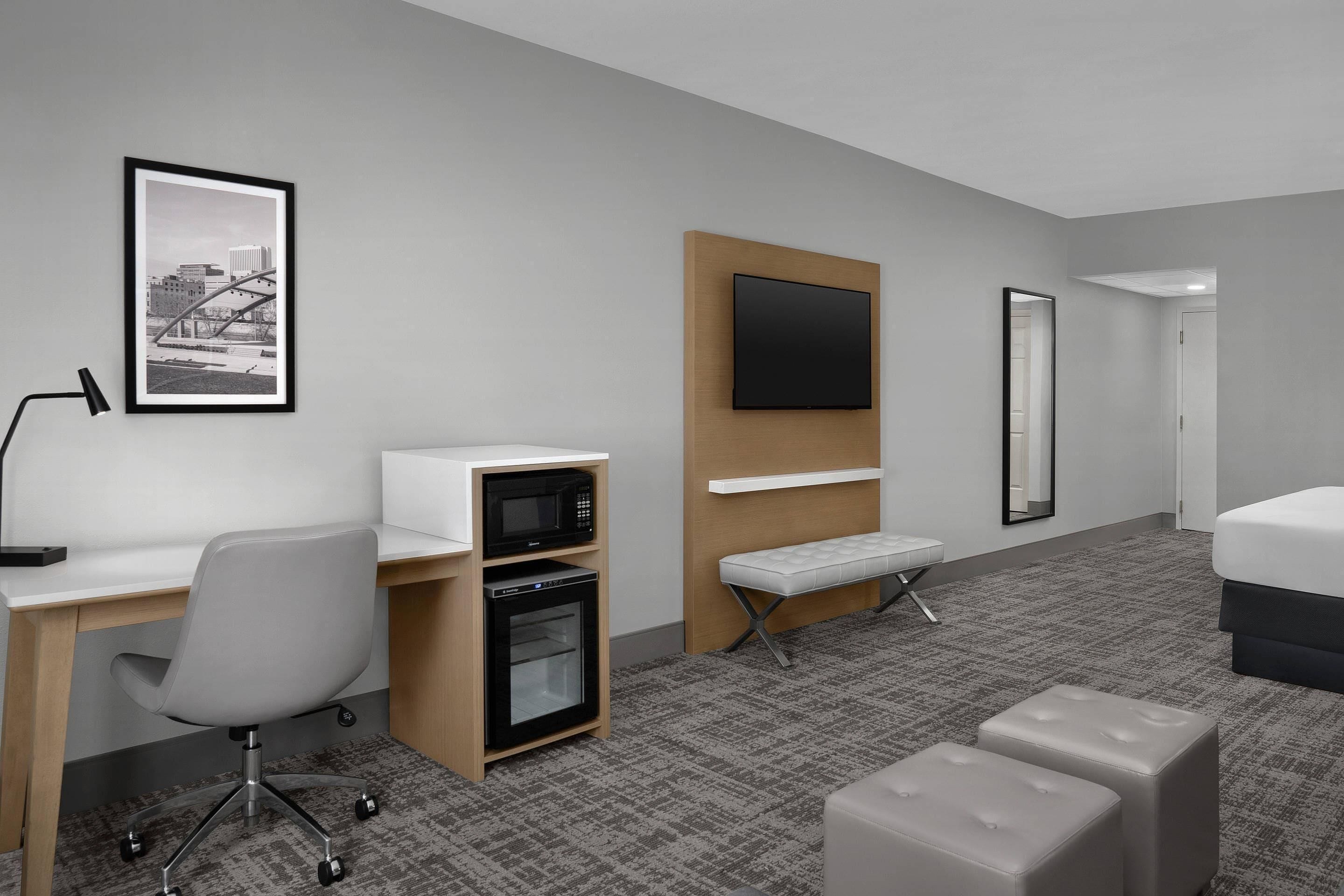 A hotel room with a desk, microwave, oven, bench, ottoman, bed, TV, and a framed picture on the wall.