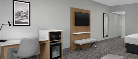 A hotel room with a desk, microwave, oven, bench, ottoman, bed, TV, and a framed picture on the wall.