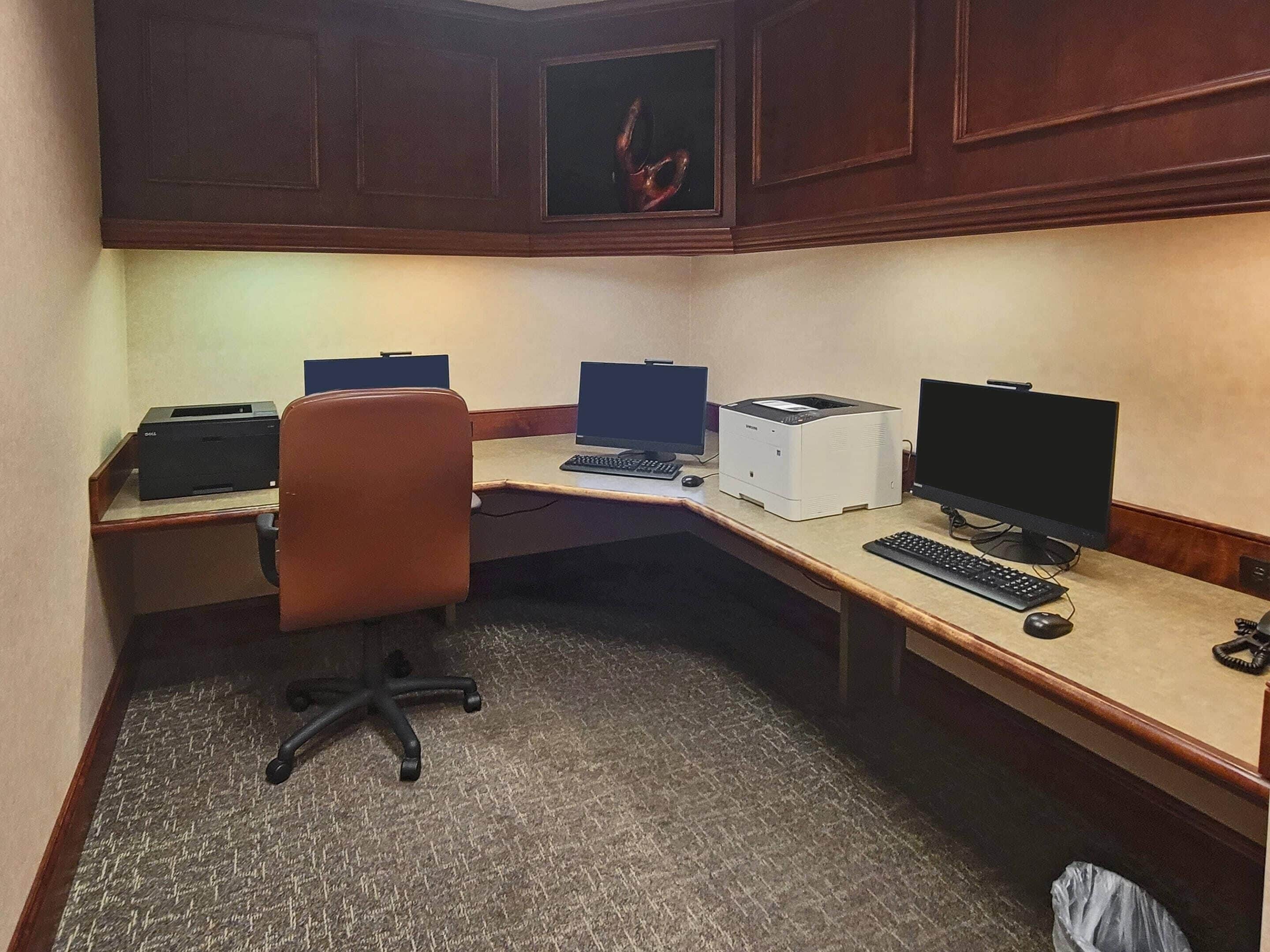 A corner office with a desk, two computer monitors, a printer, and a chair.
