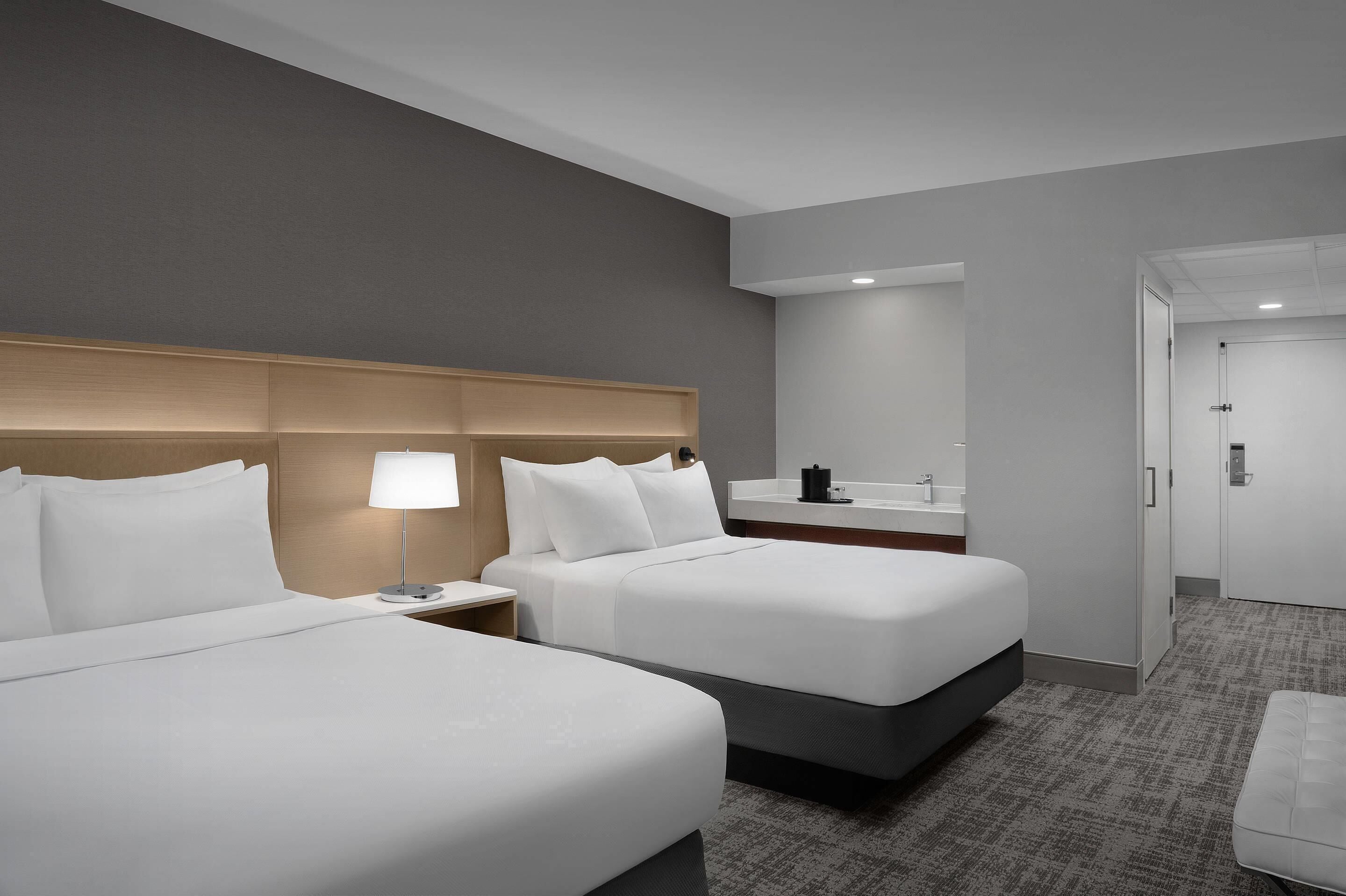 Habitación de hotel con dos camas, una mesita de noche con lámpara, un escritorio pequeño con espejo y una puerta.
