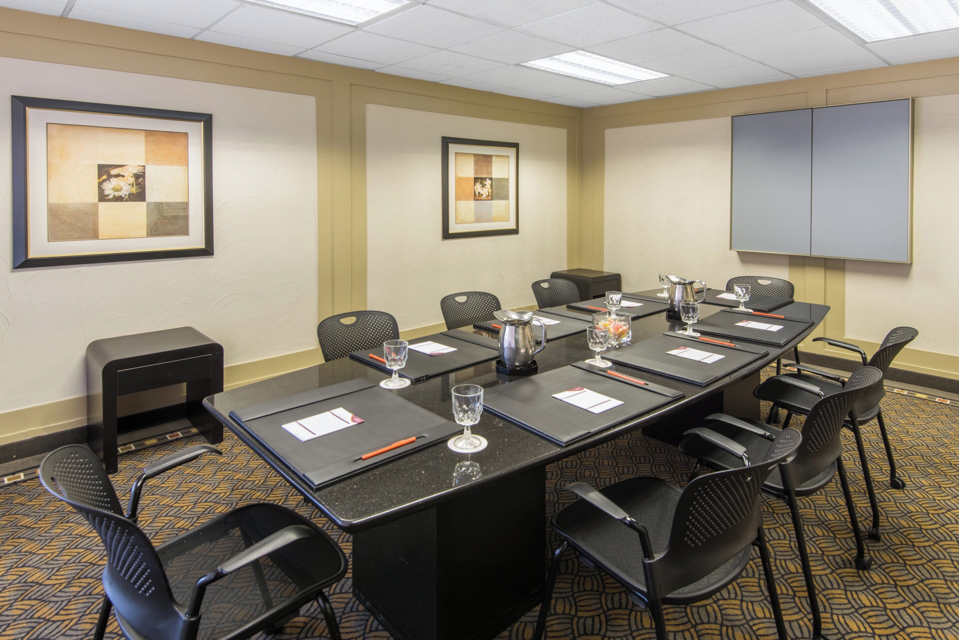 A conference room with a long table, chairs, a projector screen, and framed artwork on the walls.