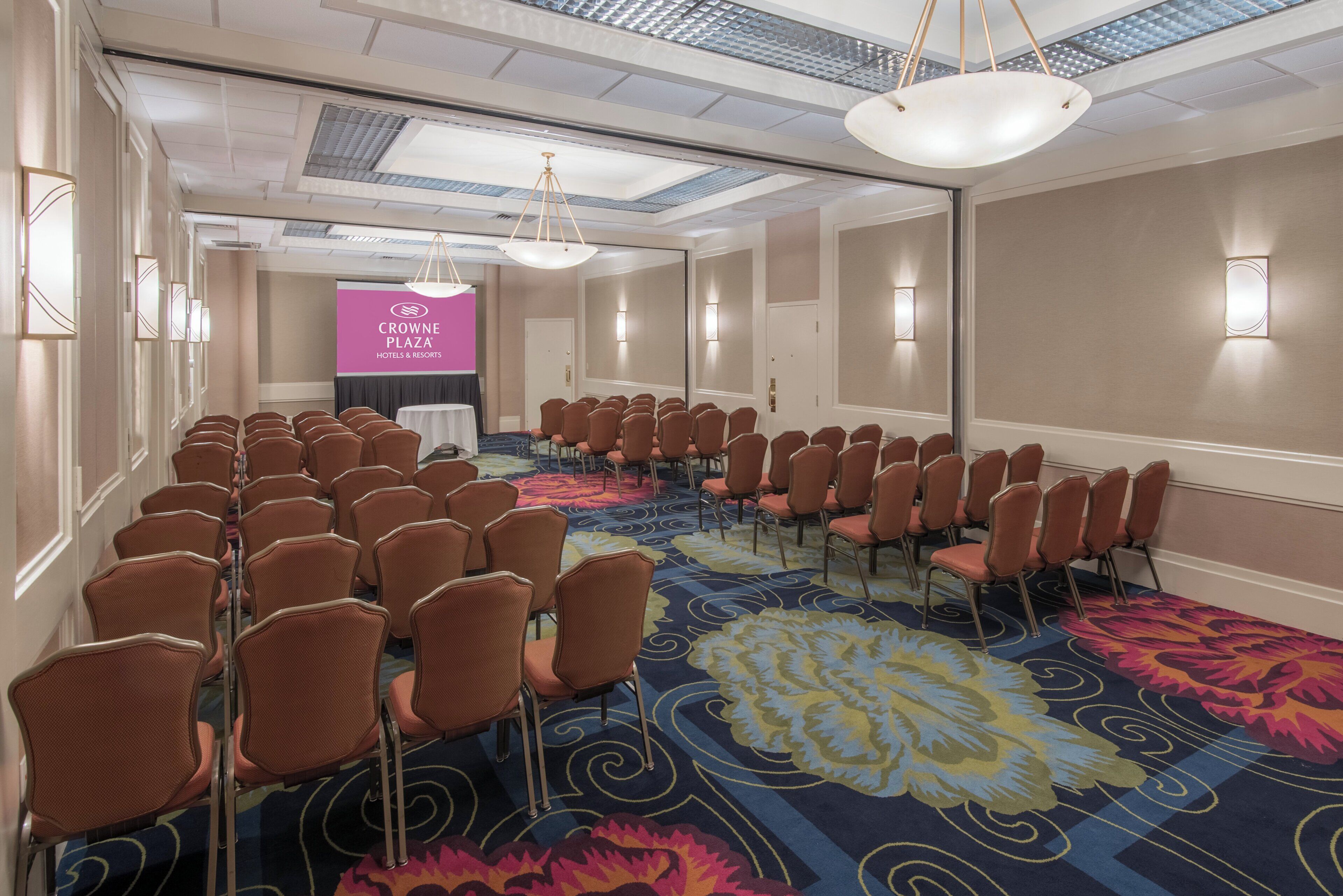 A conference room with rows of chairs, a podium, and a banner for Crowne Plaza Hotels & Resorts.