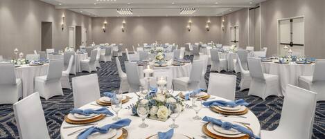 A large banquet hall with round tables set for dining, white chairs, and chandeliers.