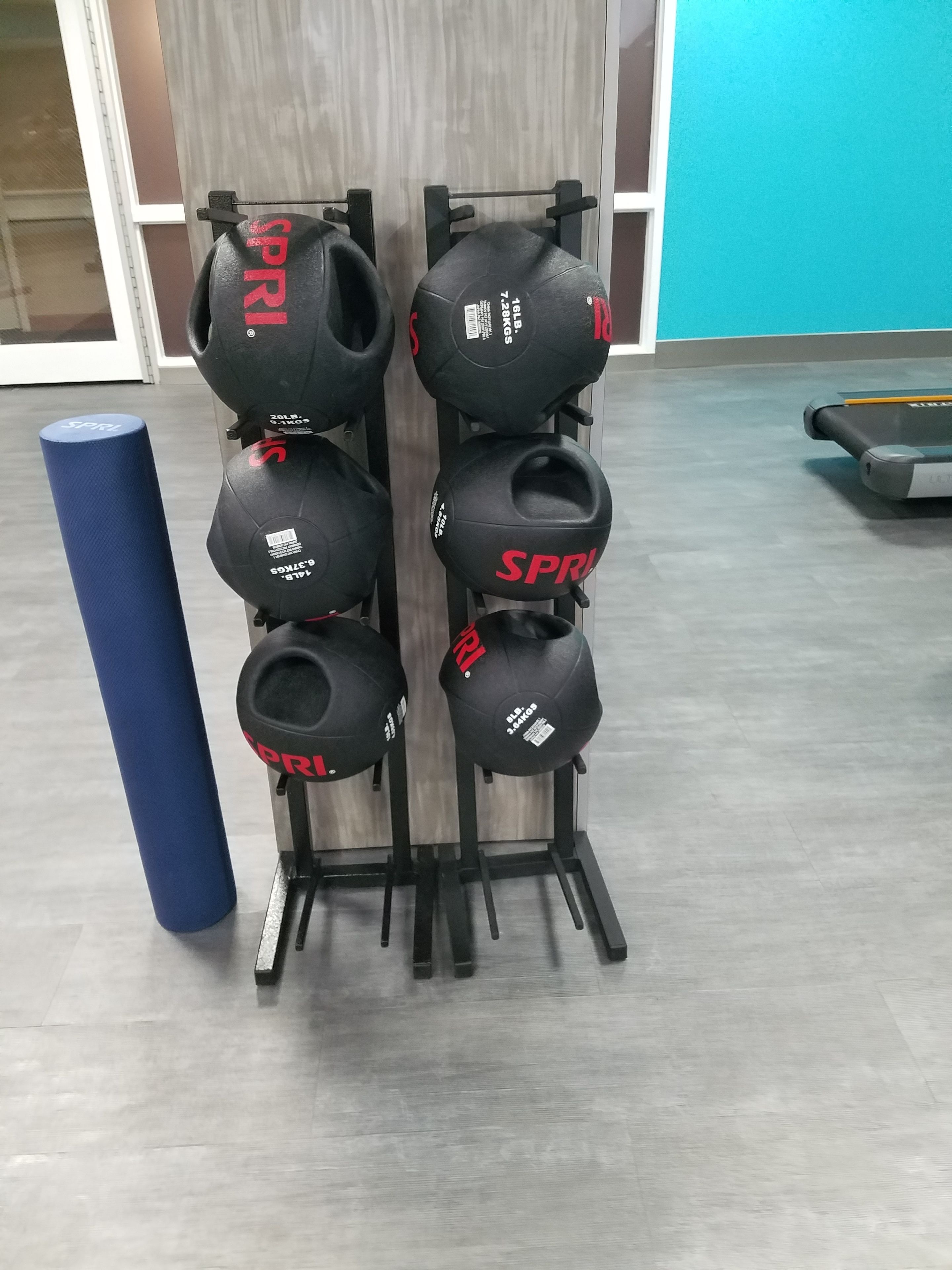 Une salle de sport avec un support mural où sont rangés différents ballons d’exercice et un tapis bleu.