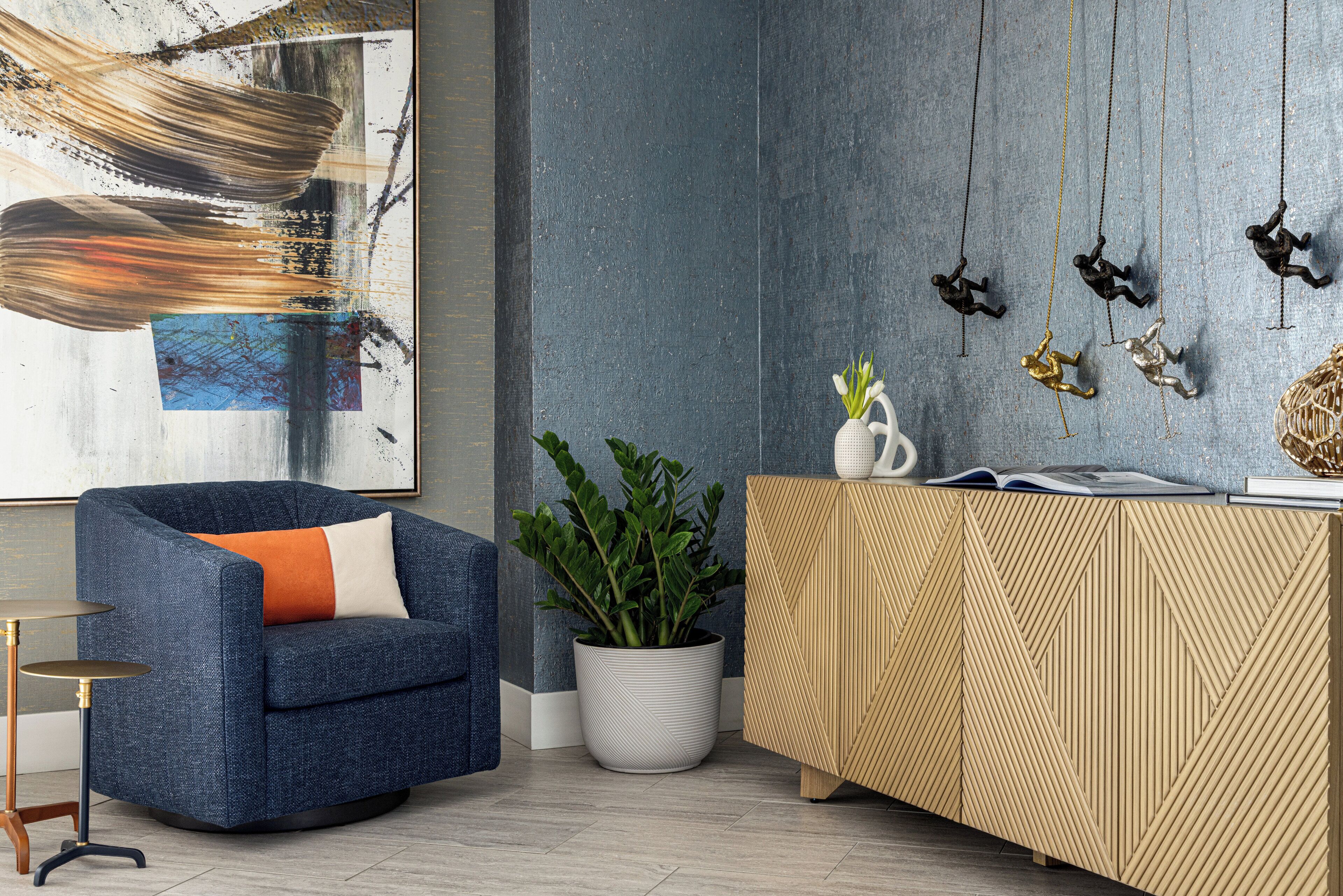 A modern living room with a blue armchair, a potted plant, a wooden cabinet, and a wall with abstract art and hanging ornaments.