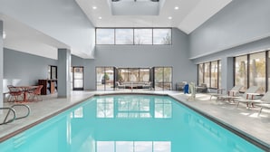 Piscina interior moderna com paredes de vidro transparente, teto alto e área de estar com cadeiras e mesa.