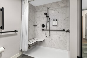 A modern bathroom with a glass-enclosed shower, marble walls, a white bench, and toiletries on a wall-mounted rack. - Crowne Plaza Virginia Beach Town Center by IHG (Virginia Beach)