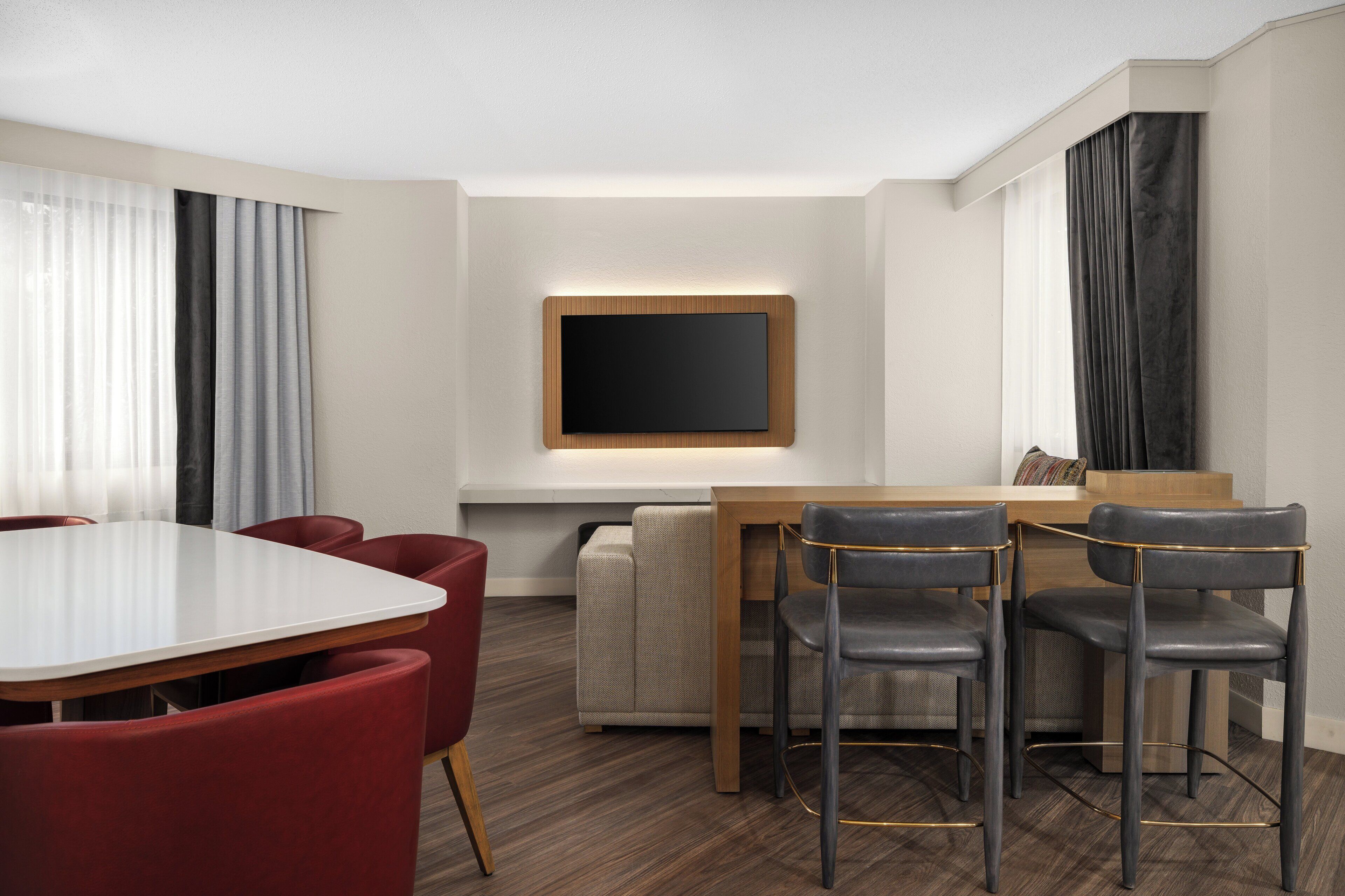 Una habitación de hotel moderna con zona de comedor que incluye una mesa blanca, sillas rojas y un sofá.