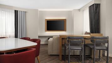 Una habitación de hotel moderna con zona de comedor que incluye una mesa blanca, sillas rojas y un sofá.