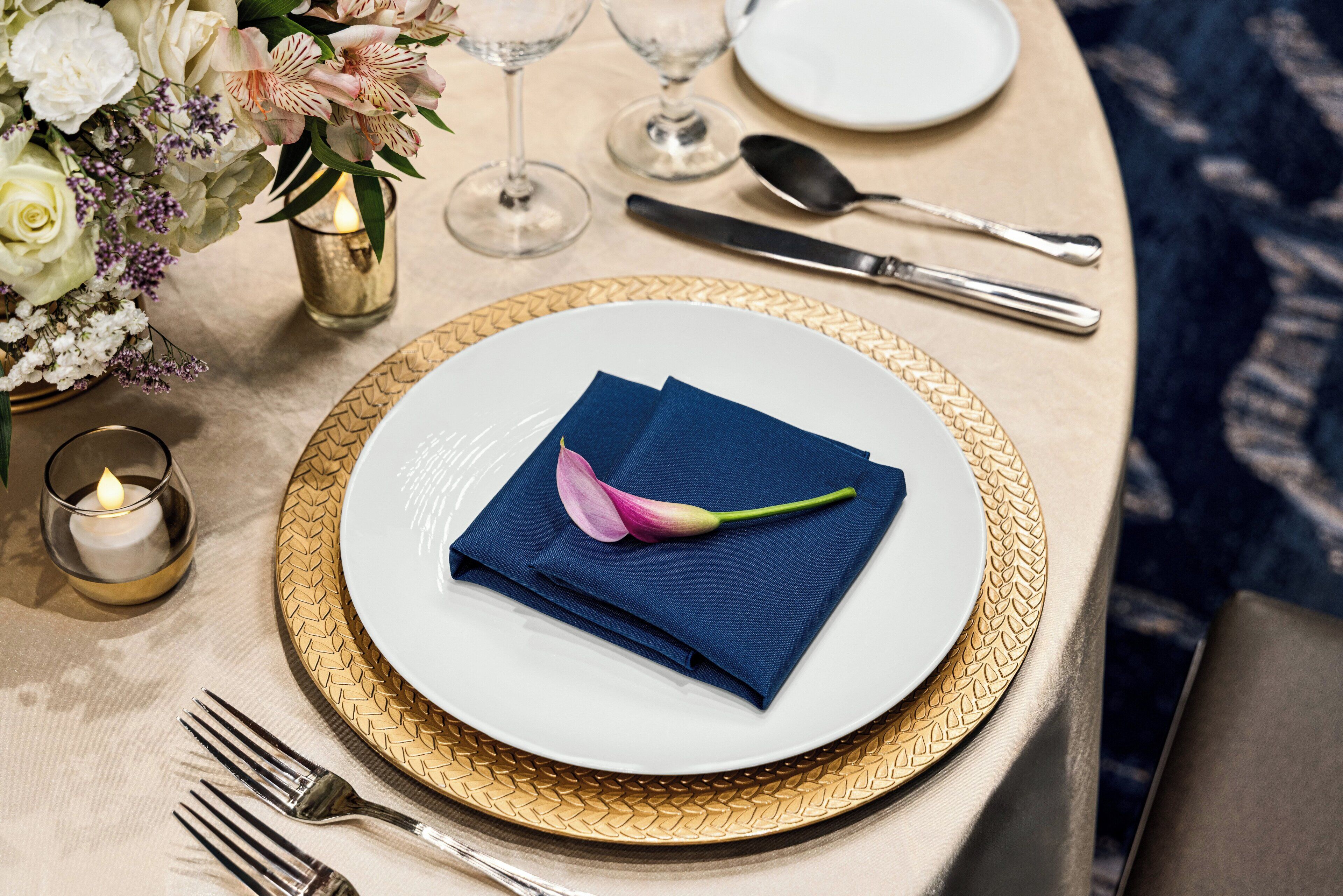 A table set with a white plate, gold rimmed charger, folded blue napkin, and a pink flower.