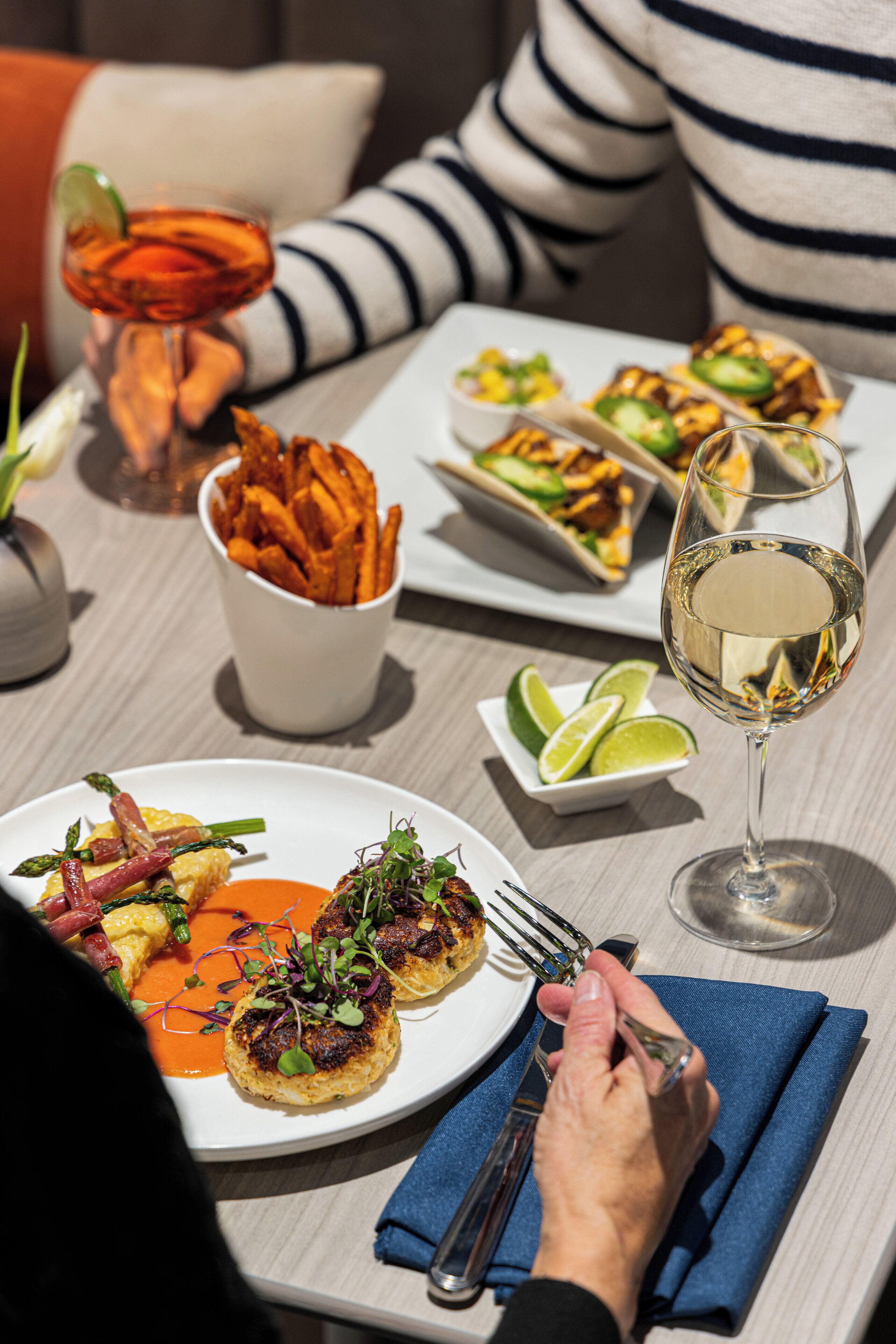 A dining table with plates of food, a glass of white wine, and a bowl of fries.