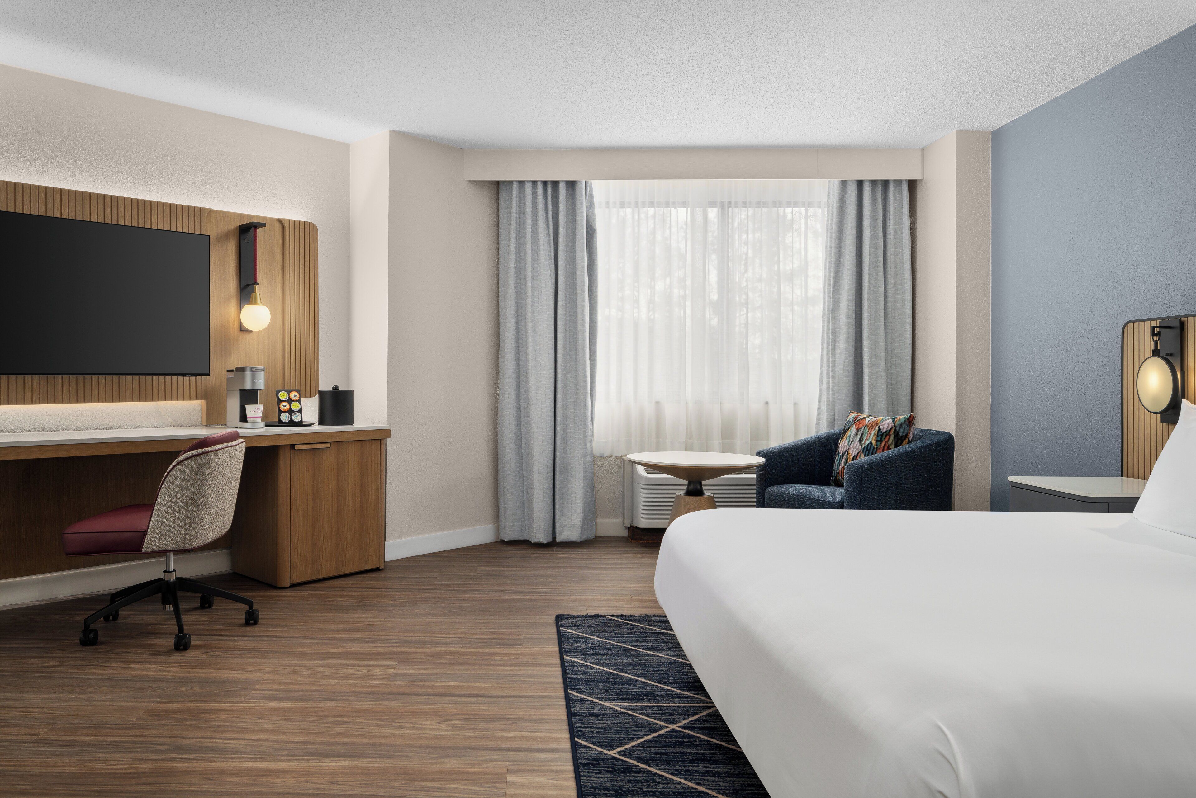 Quarto de hotel moderno com uma cama grande, uma escrivaninha com cadeira, televisão na parede, uma mesa pequena e uma janela com cortinas.