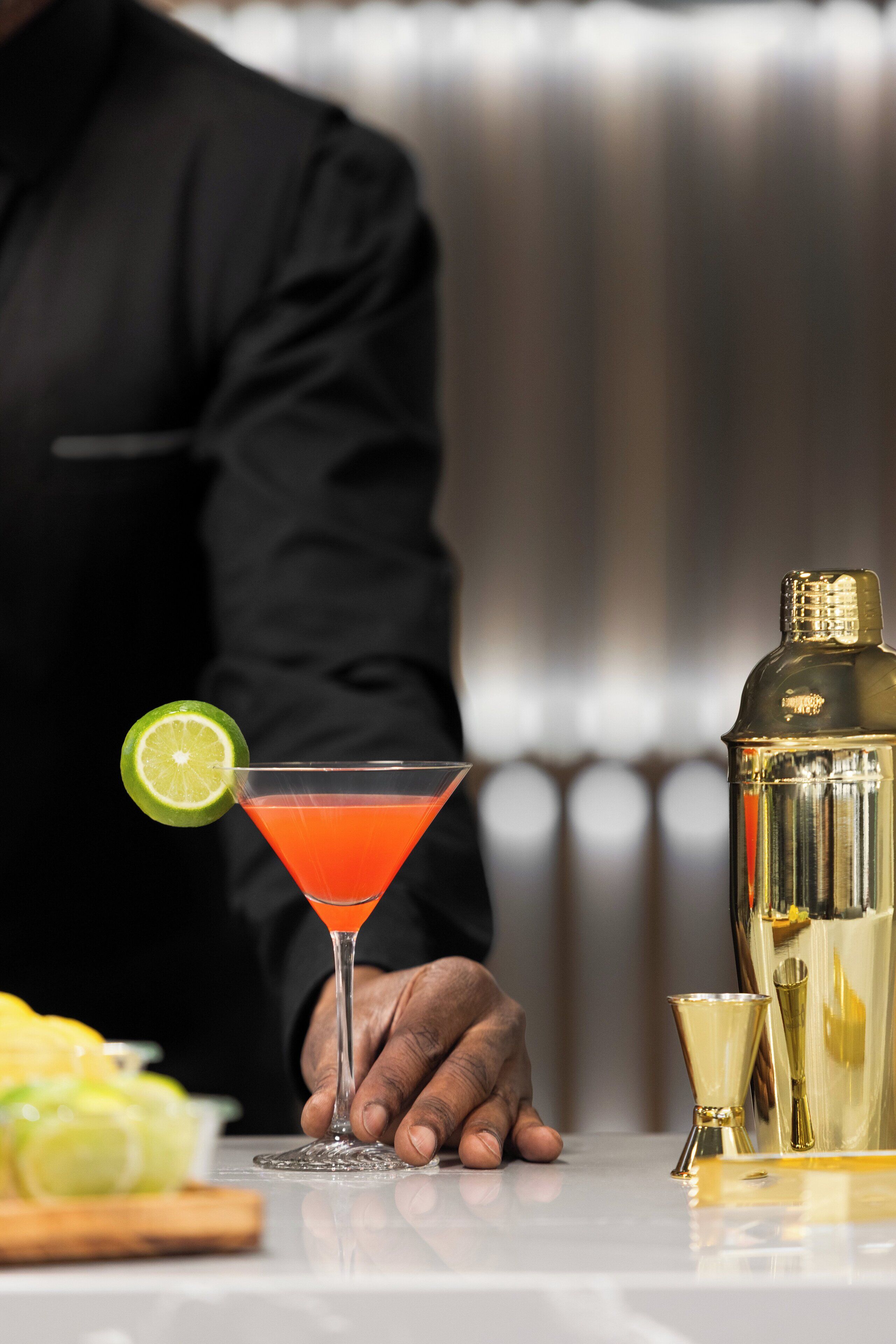 En bartender holder et martiniglas med en limeskive, en cocktailshaker og en barged på bardisken.