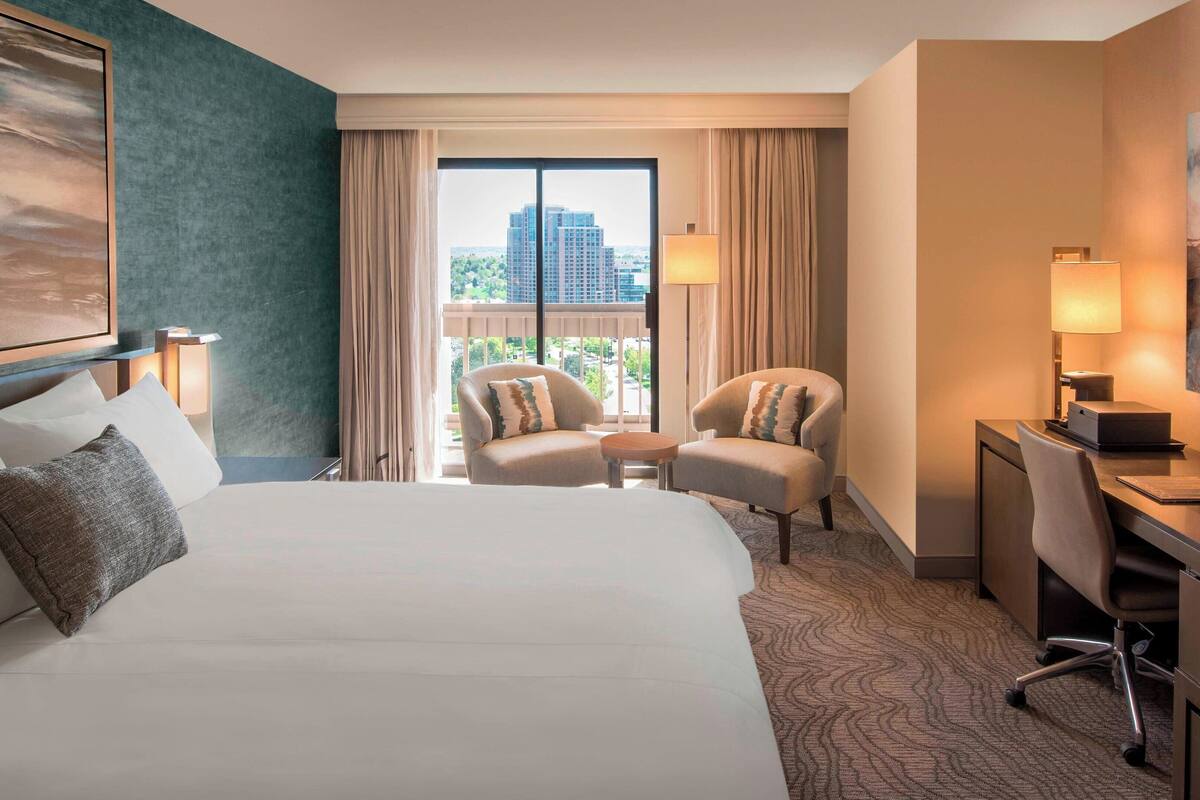 A hotel room with a large bed, two armchairs, a desk with a chair, and a view of a cityscape through the window.