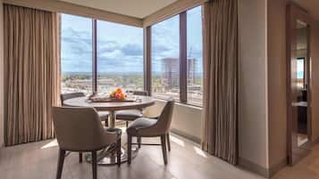 A modern hotel room with a large window offering a city view, a round dining table with chairs, and a visible balcony.