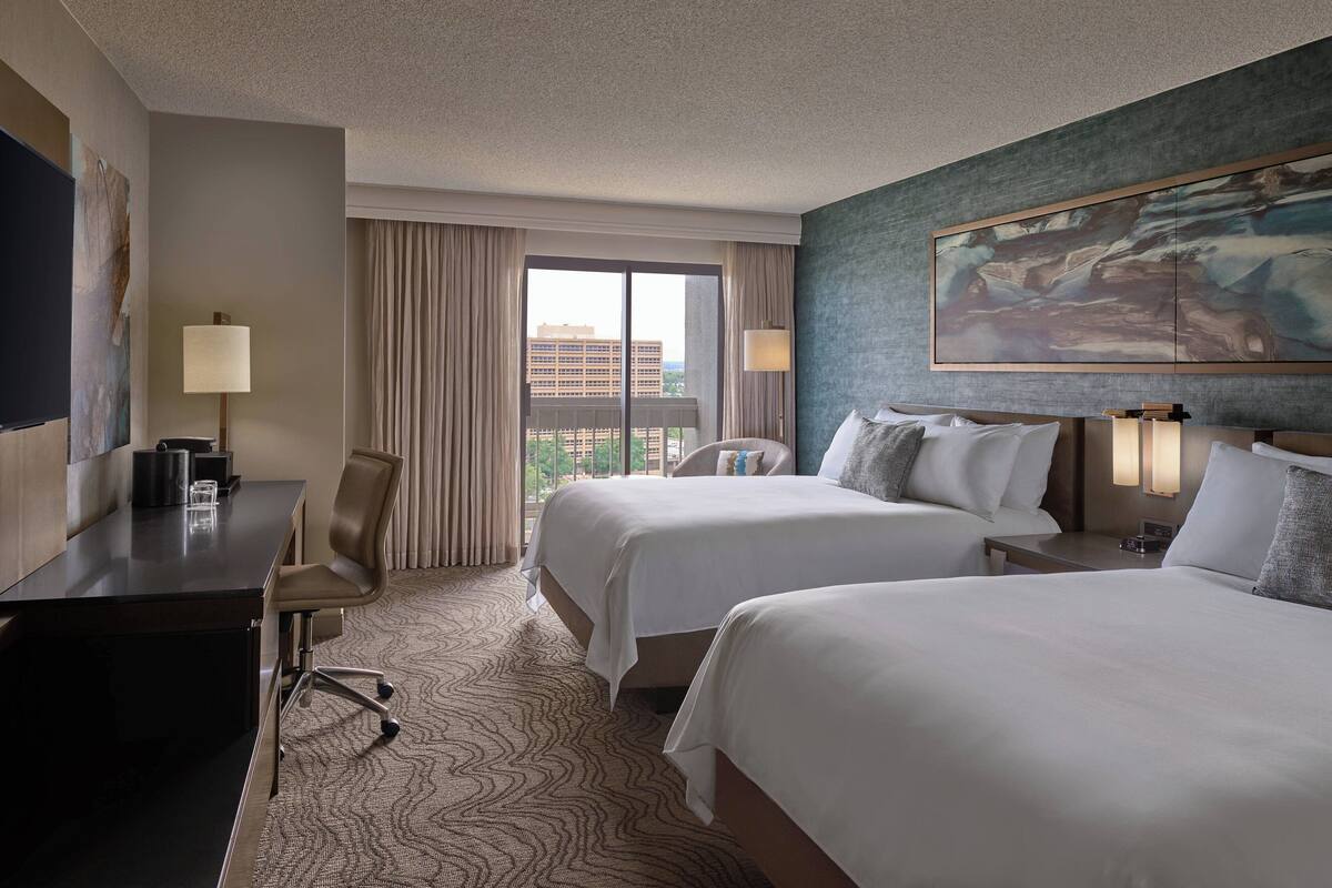 A hotel room with two beds, a desk, a chair, a large window, and a painting on the wall.