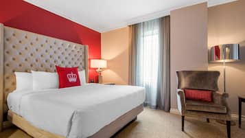 Une chambre d’hôtel avec un grand lit, un oreiller rouge, une chaise, une lampe et une fenêtre avec des rideaux.