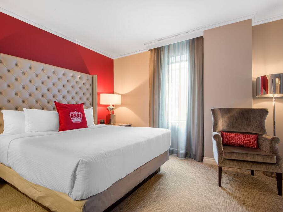 Une chambre d’hôtel avec un grand lit, un oreiller rouge, une chaise, une lampe et une fenêtre avec des rideaux.