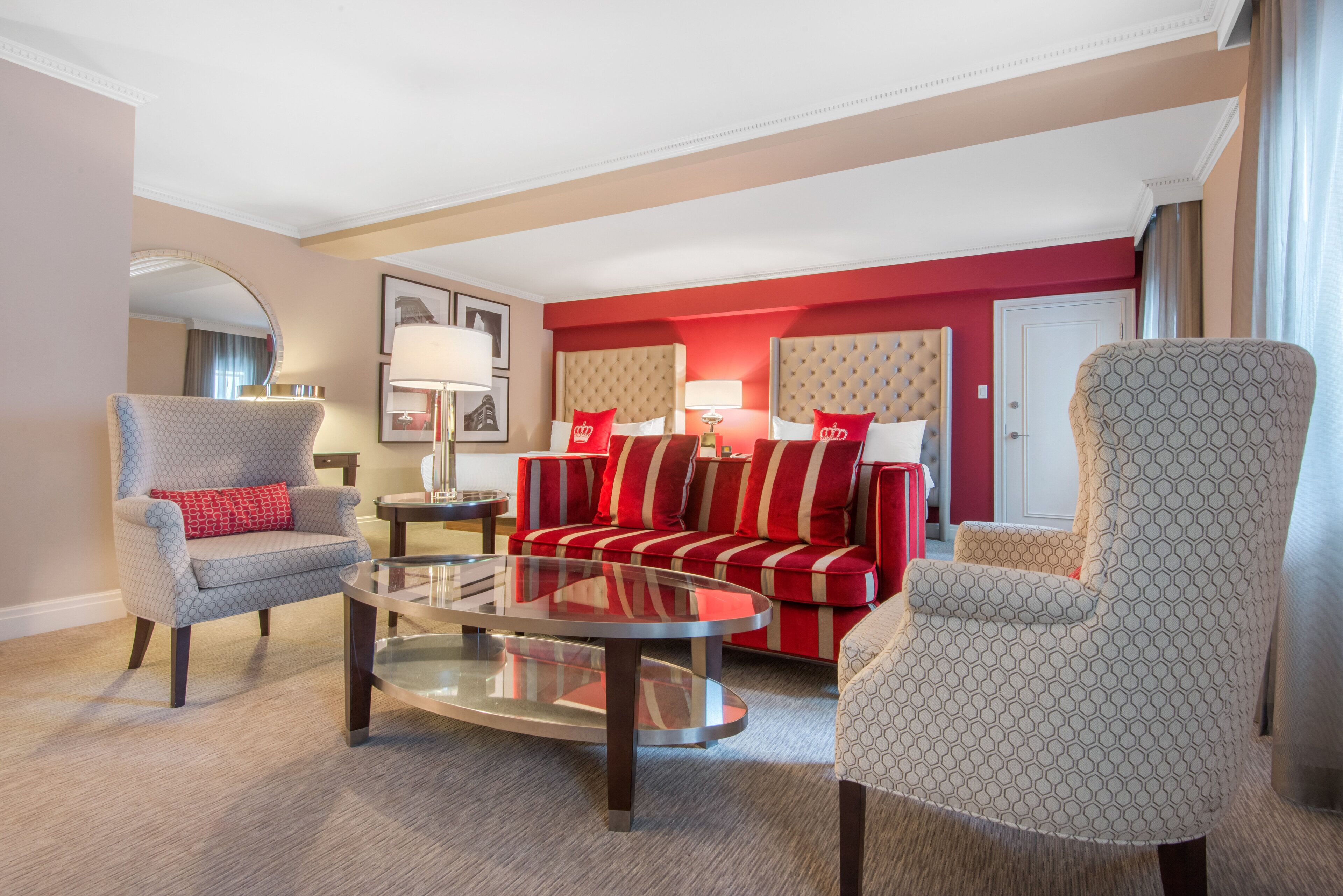 A modern hotel room with a red and beige color scheme, featuring a sofa, armchairs, a glass coffee table, and a large mirror.