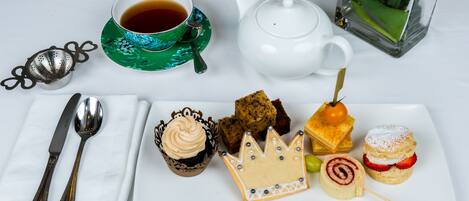 A tea set with a teapot and cups, a sugar bowl, a creamer, a cupcake, a slice of cake, a sandwich, and a dessert on a white plate.