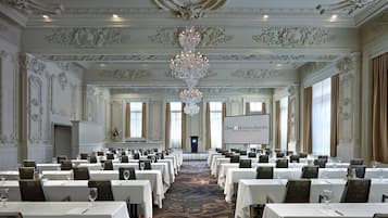 Un salón de conferencias con detalles ornamentales en el techo, una gran lámpara de araña y una pantalla que muestra