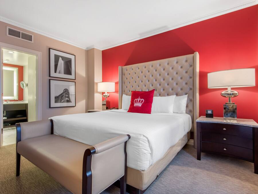 Quarto de hotel com cama grande, mesa de cabeceira, cadeira e parede com detalhe em vermelho.