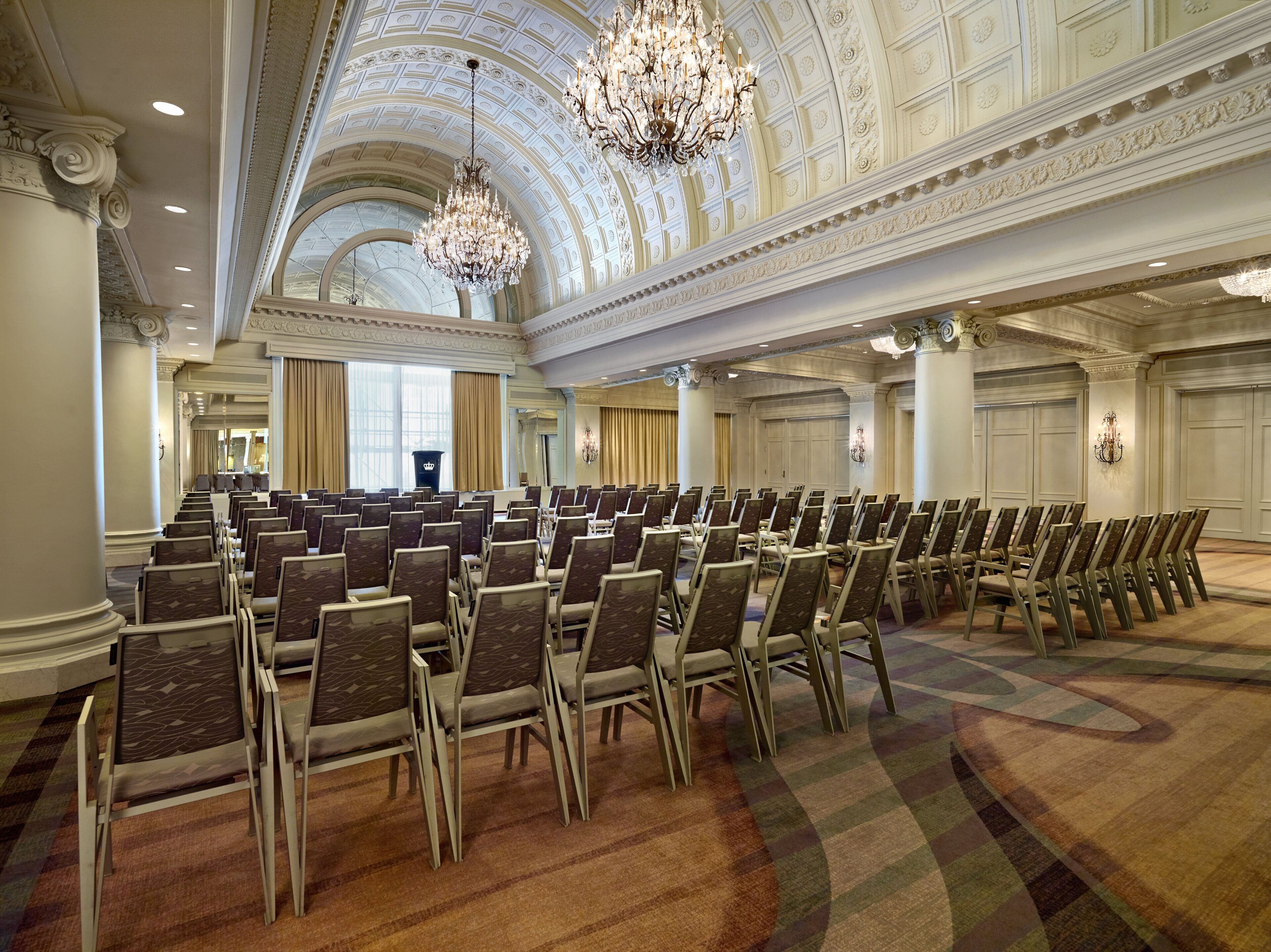 A large conference room with rows of chairs, a high ornate ceiling, chandeliers, and large windows.
