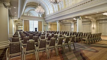 A large conference room with rows of chairs, a high ornate ceiling, chandeliers, and large windows.