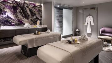 A modern spa room with two massage tables, a large abstract painting, a robe hanging on the wall, and a small round table with a bottle and a glass.