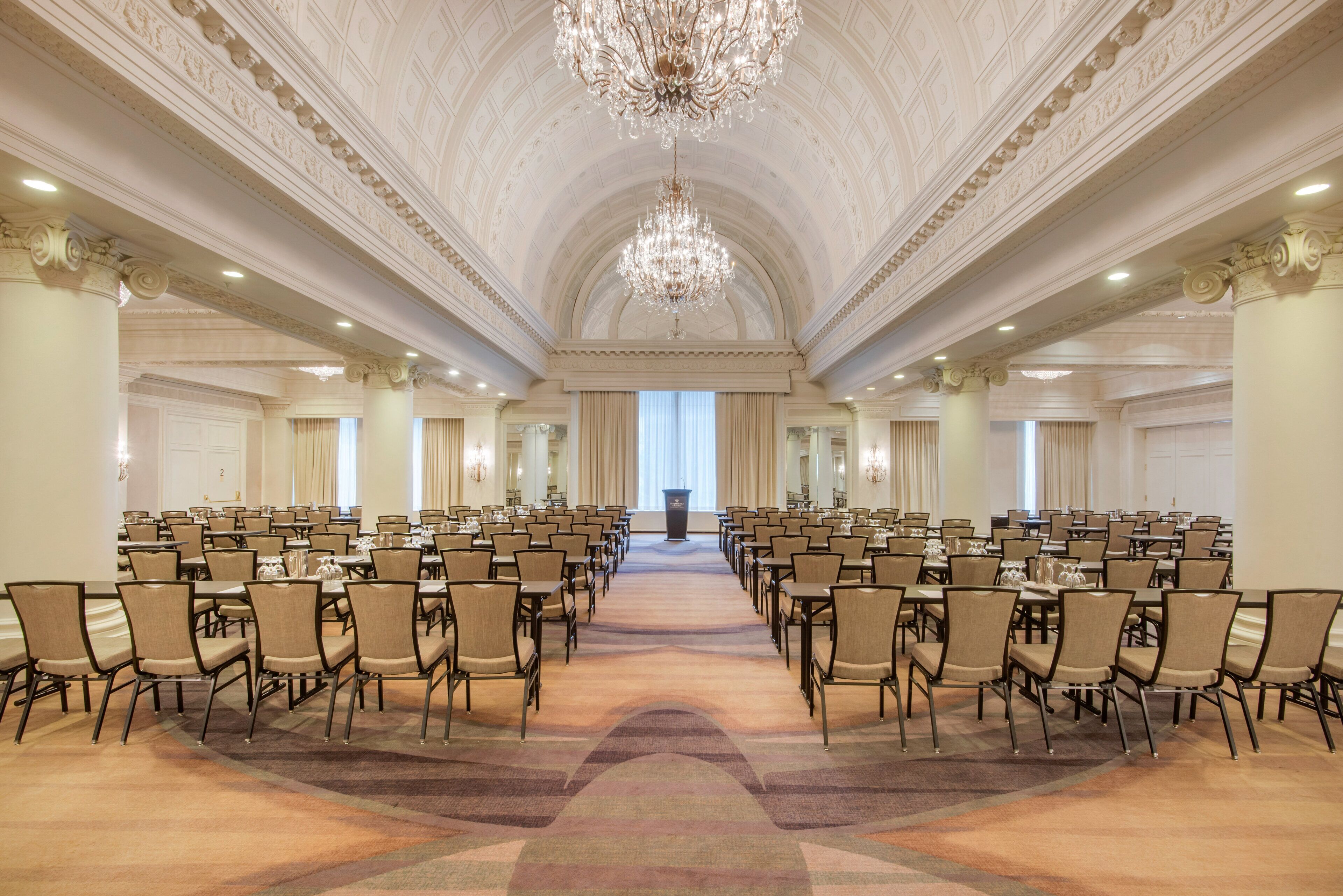 A large conference room with rows of tables and chairs, a grand chandelier, and a high ceiling with detailed moldings.