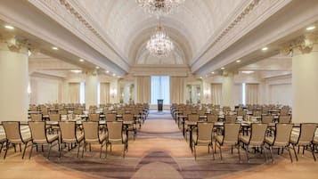 A large conference room with rows of tables and chairs, a grand chandelier, and a high ceiling with detailed moldings.