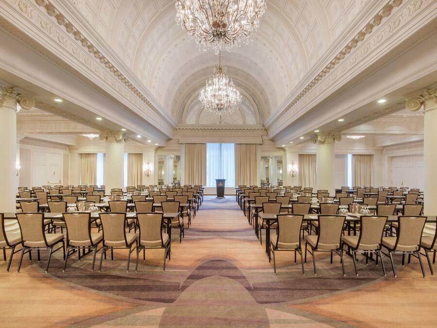 A large conference room with rows of tables and chairs, a grand chandelier, and a high ceiling with detailed moldings.