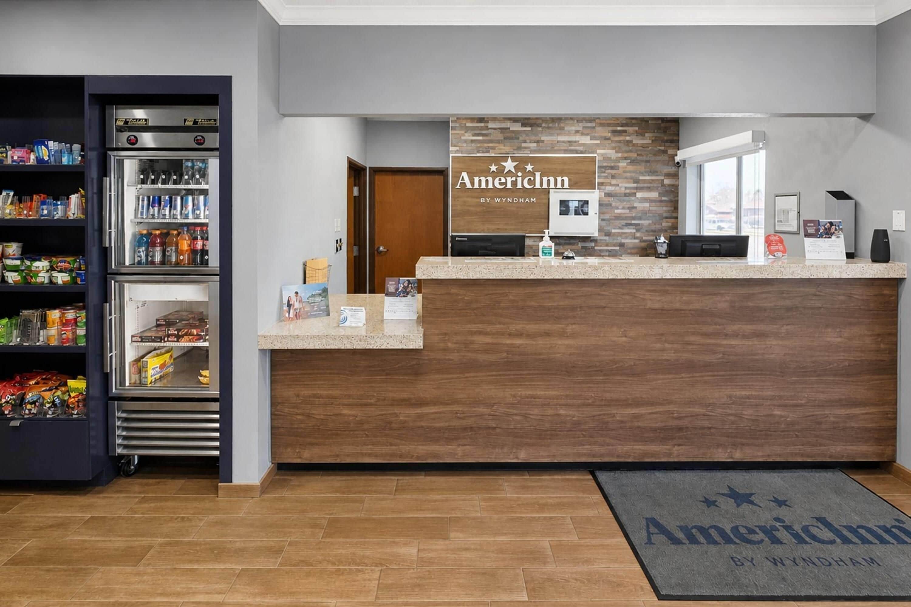 Una reception con una insegna per AmericInn by Wyndham, un frigorifero fornito di bevande e un tappetino d'ingresso con il nome dell'hotel.