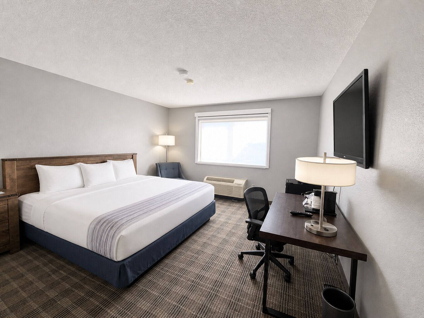 A hotel room with a bed, desk, chair, lamp, and a TV on the wall.