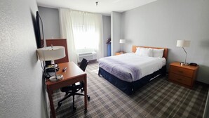 A hotel room with a bed, a desk with a chair, a bedside table with a lamp, and a window with curtains. - AmericInn by Wyndham Minot (Minot)
