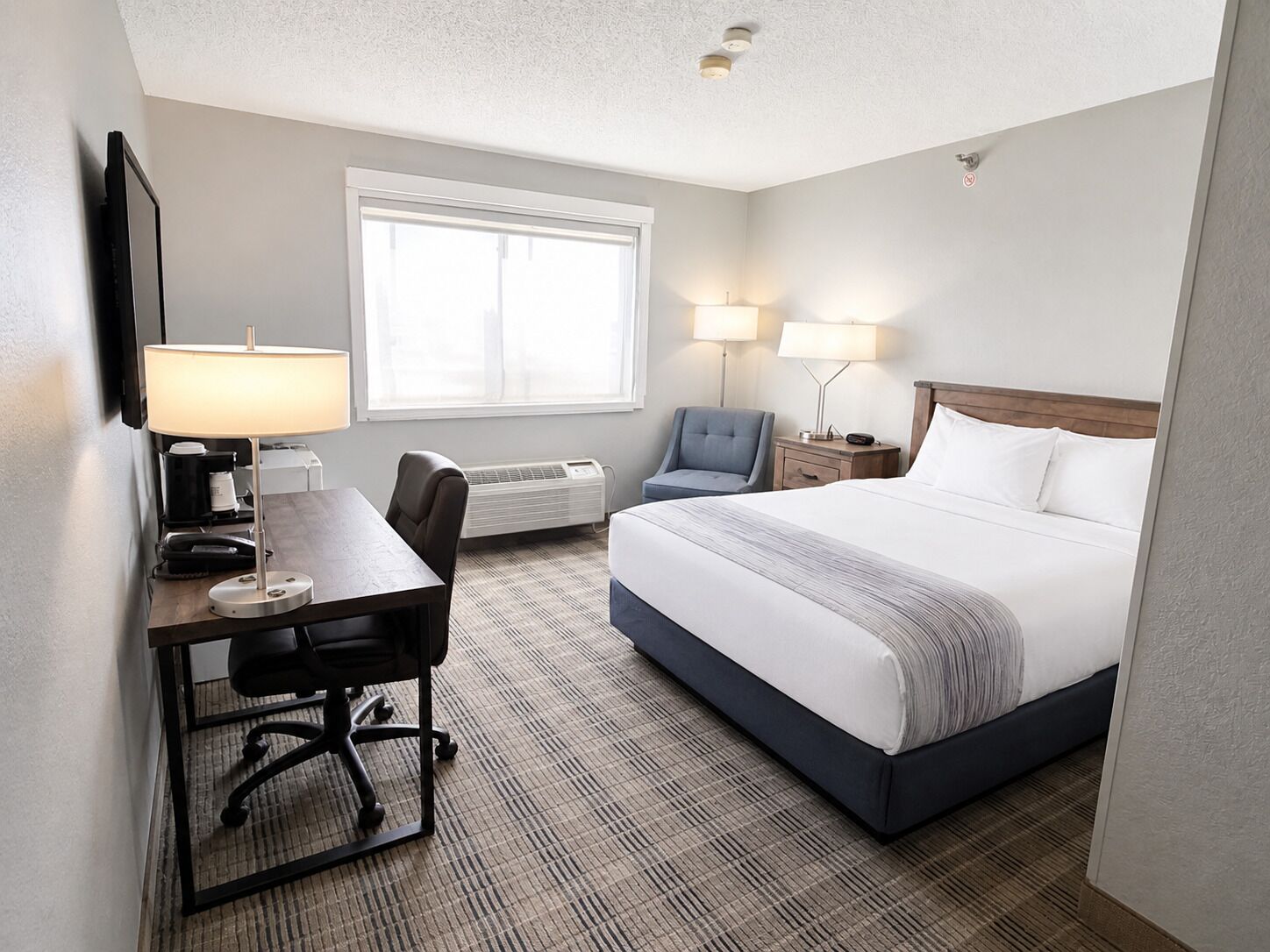 A hotel room with a bed, desk, chair, lamps, and a window.