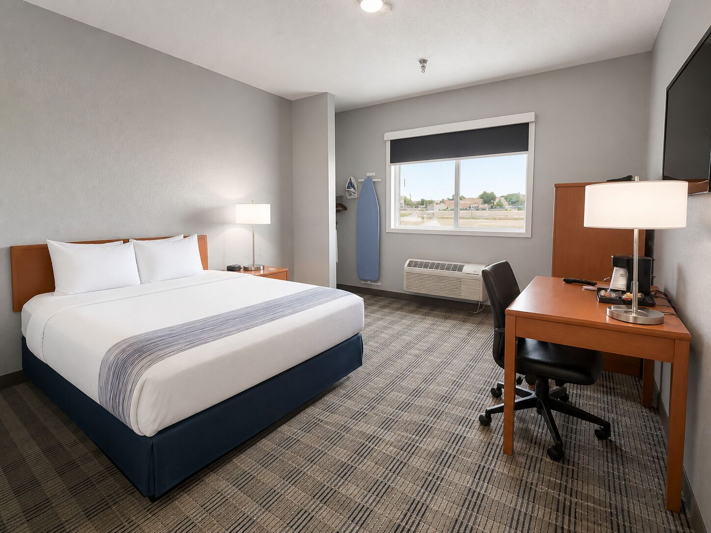 A hotel room with a bed, a desk, a chair, a lamp, a window, and a wall-mounted surfboard.