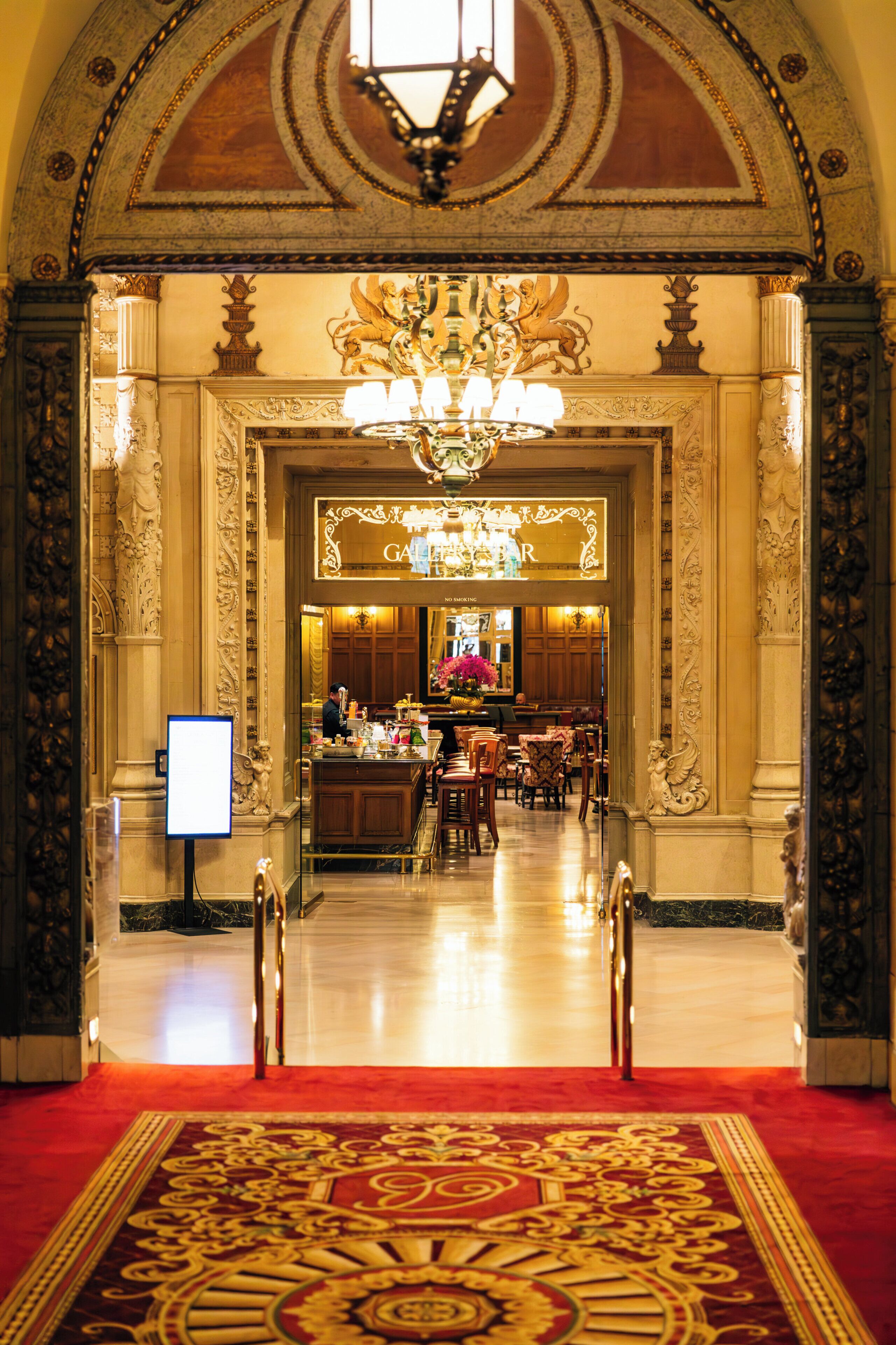 An ornate hotel lobby with a grand chandelier, a red carpet, and a bar area.