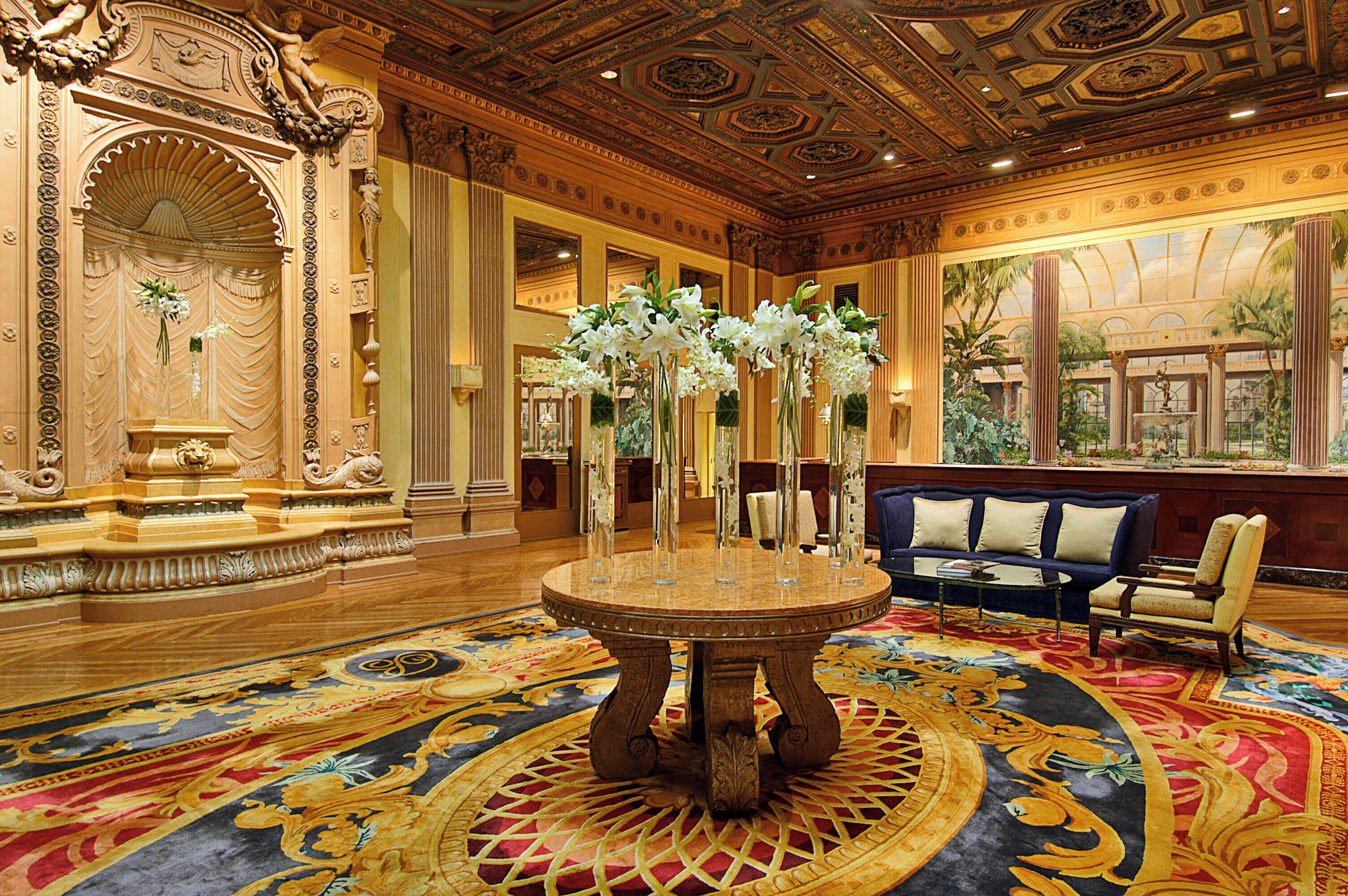 A grand lobby with ornate columns, a central round table with vases, and a blue sofa.