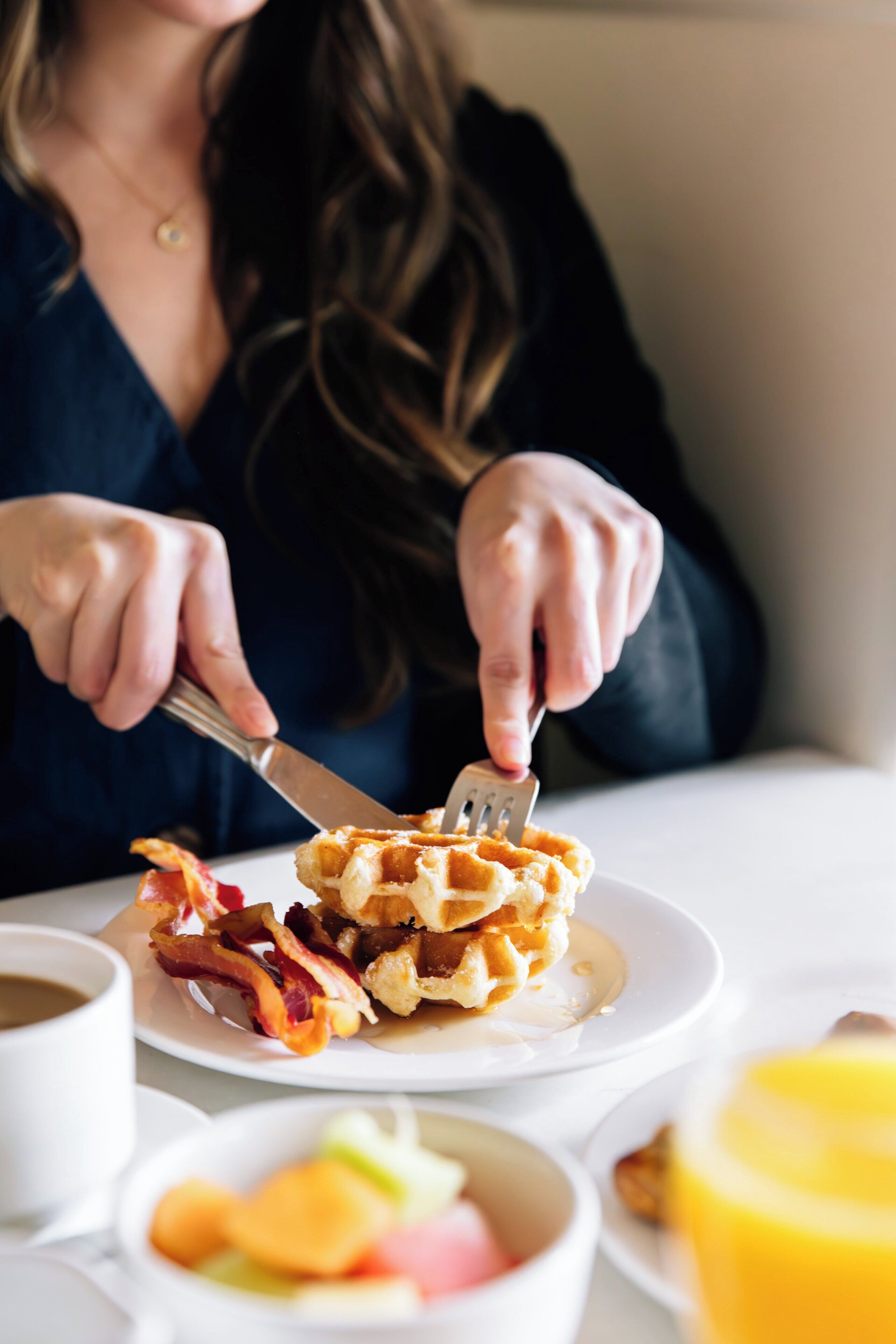 A person eating waffles with bacon and eggs, with a cup of coffee and a fruit bowl on the table.
