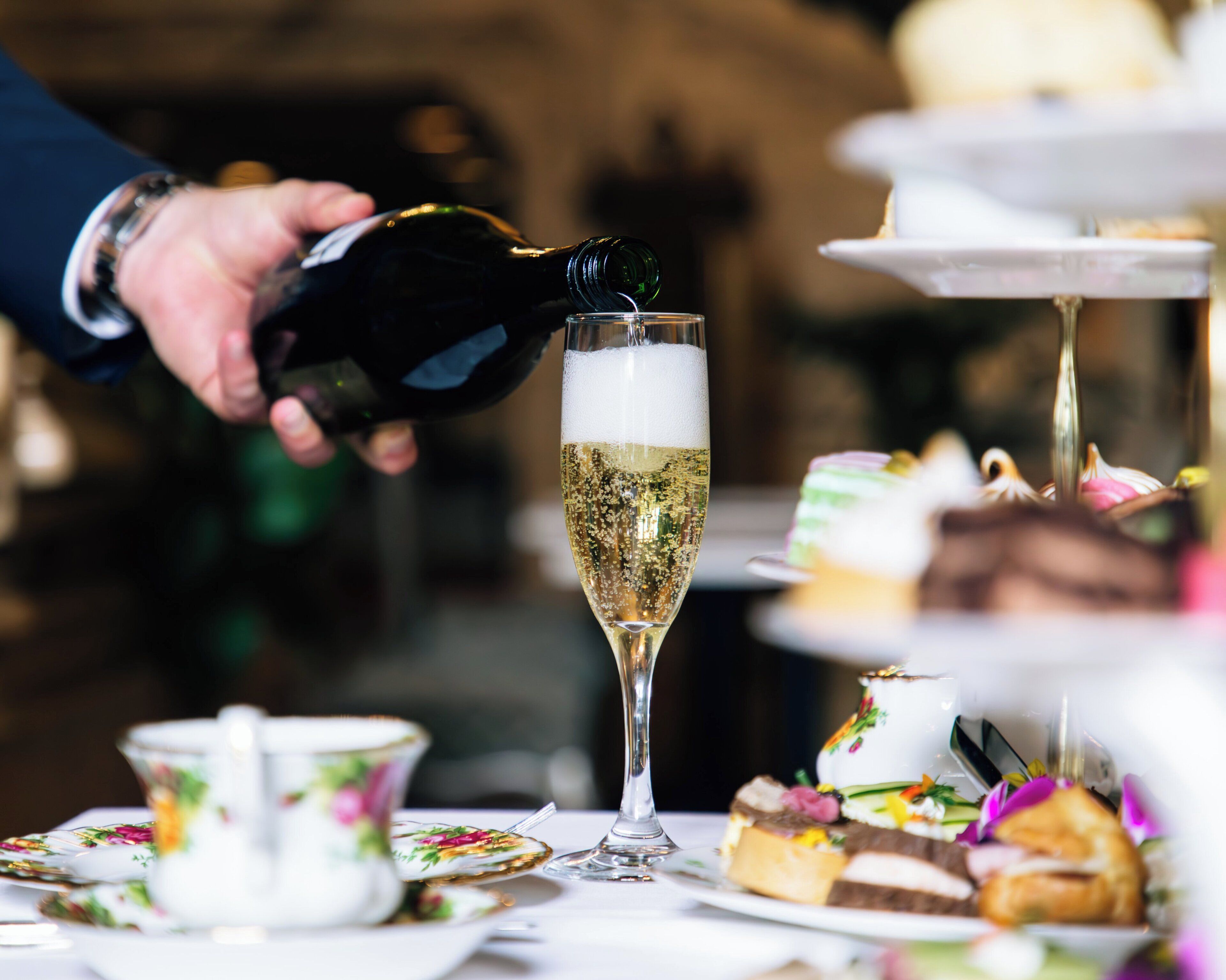 A hand pouring champagne into a glass, surrounded by a table with floral-patterned tea cups and a tiered dessert display.
