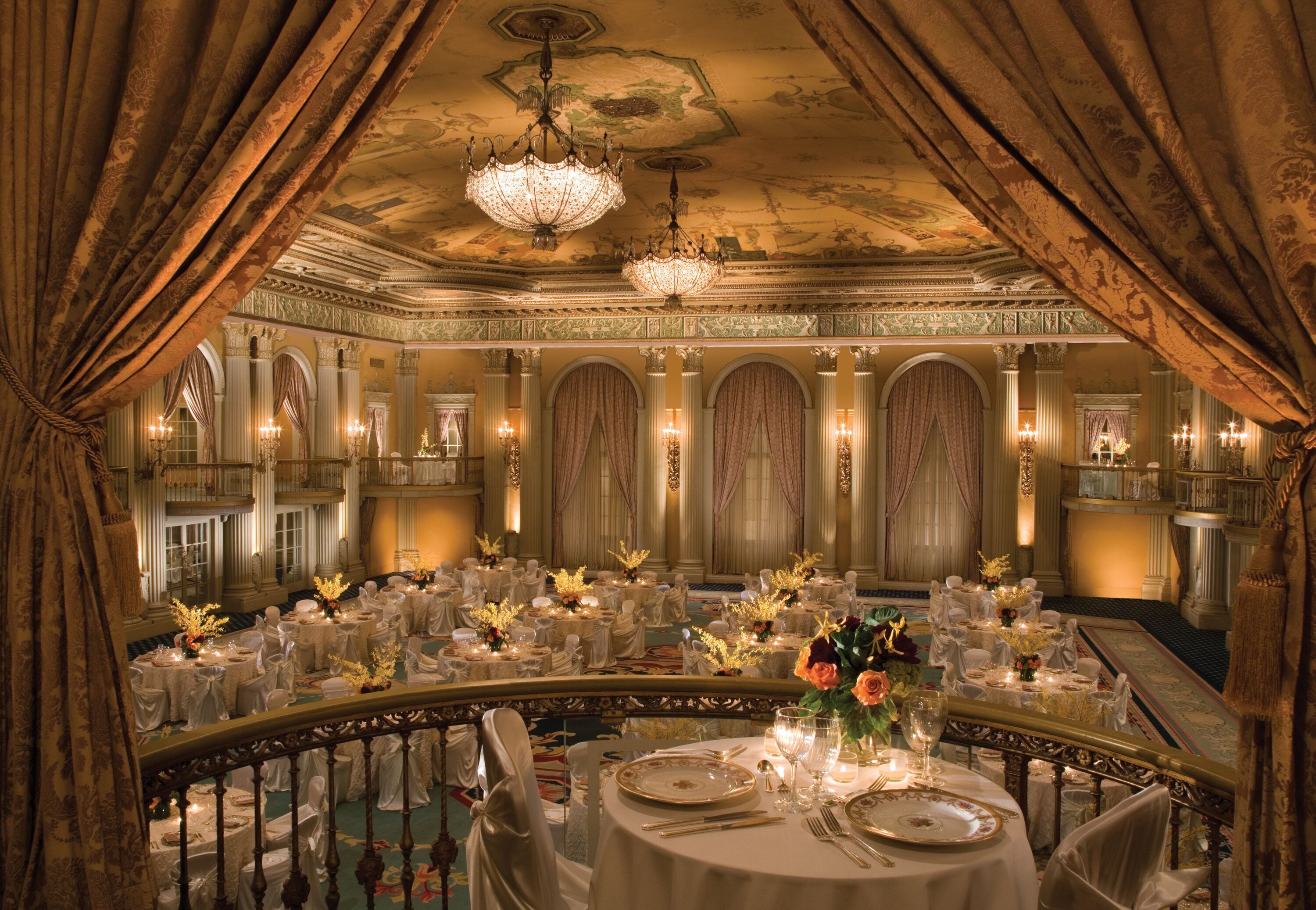 A grand ballroom with ornate chandeliers, elegant round tables, and a balcony overlooking the room.