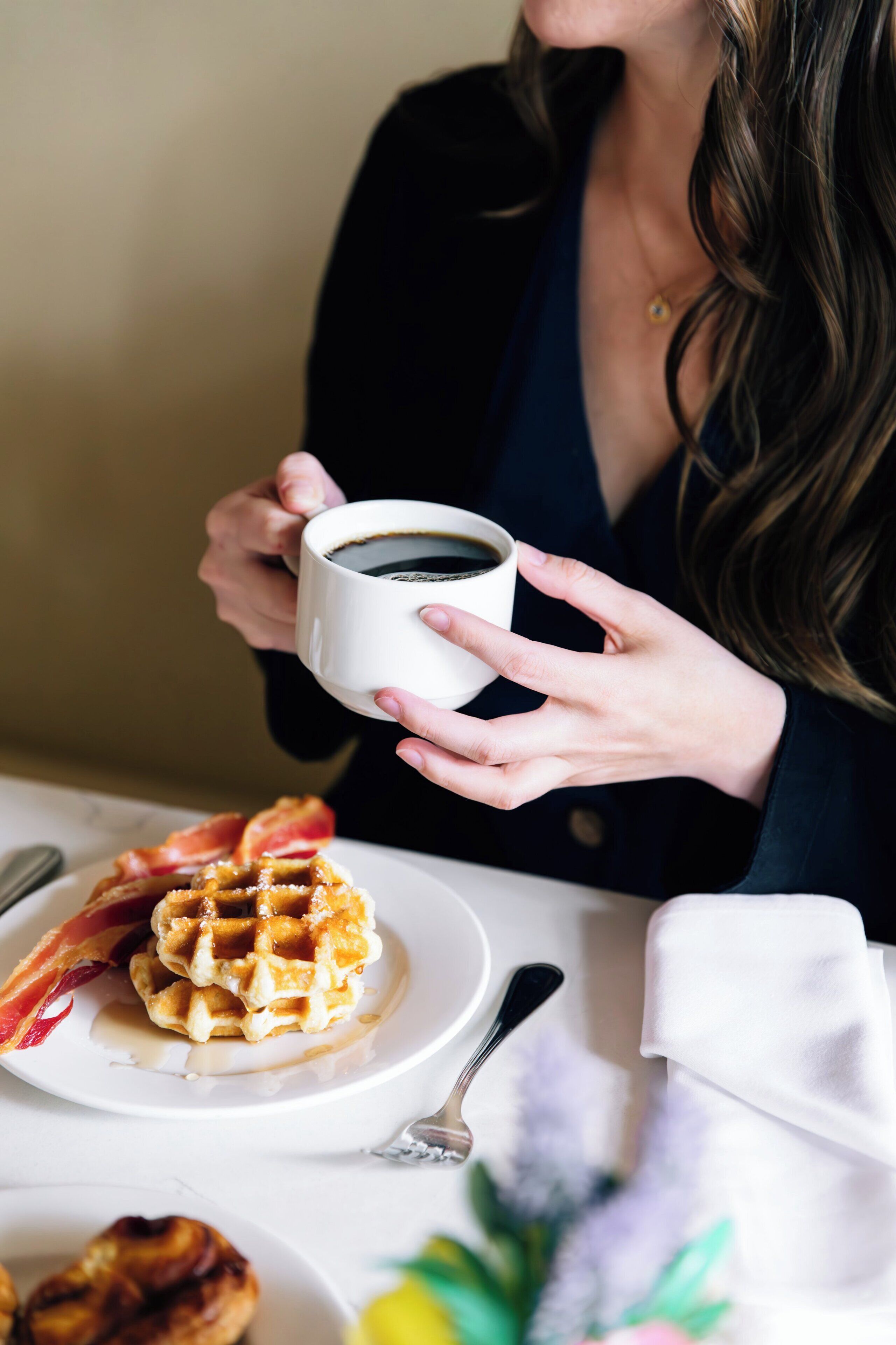Una persona tiene in mano una tazza di caffè, un piatto con waffle e pancetta, e un cucchiaio su un tavolo.
