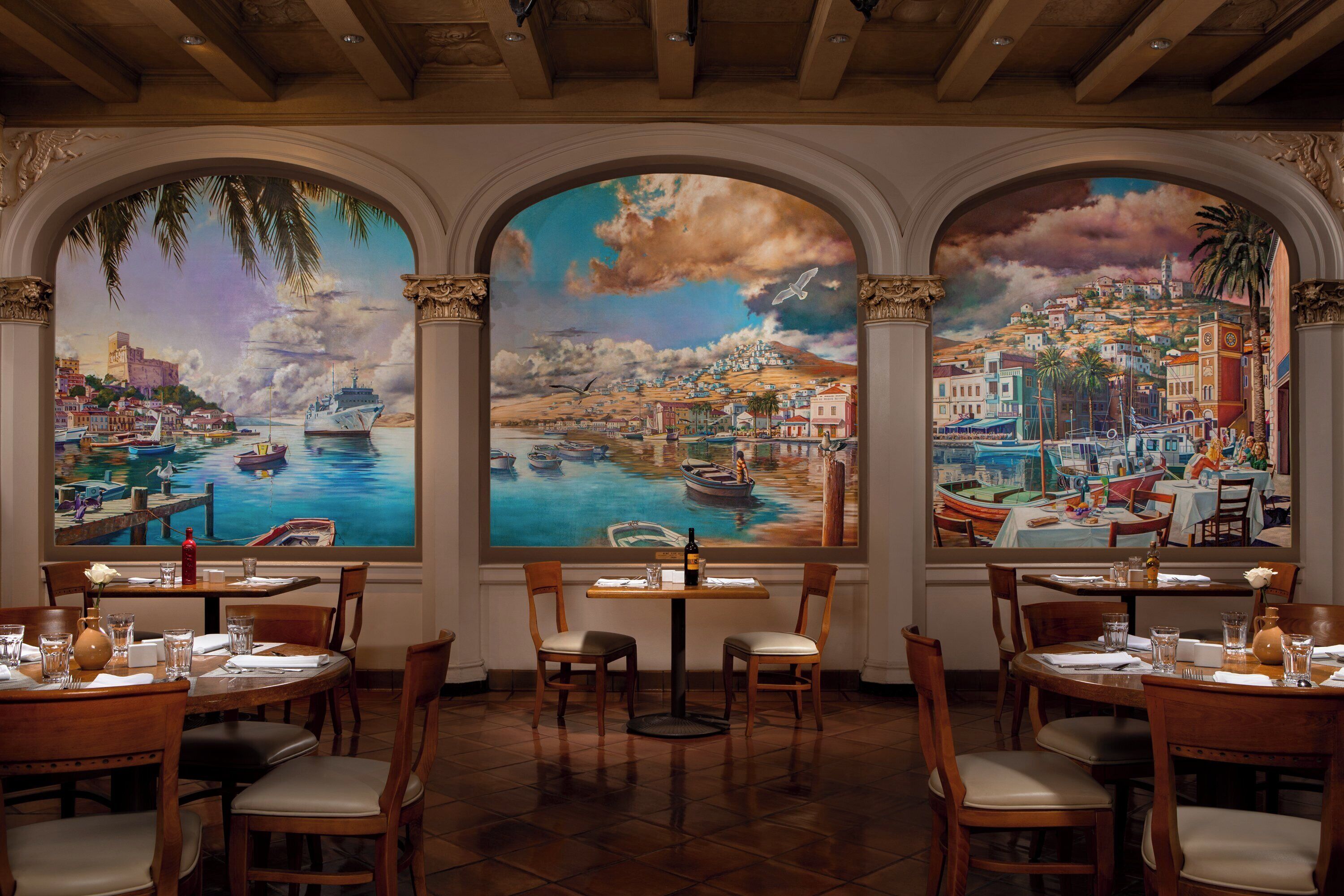 A restaurant with three large windows, each with a mural of a coastal scene. Tables are set with chairs, and there are bottles on the tables.