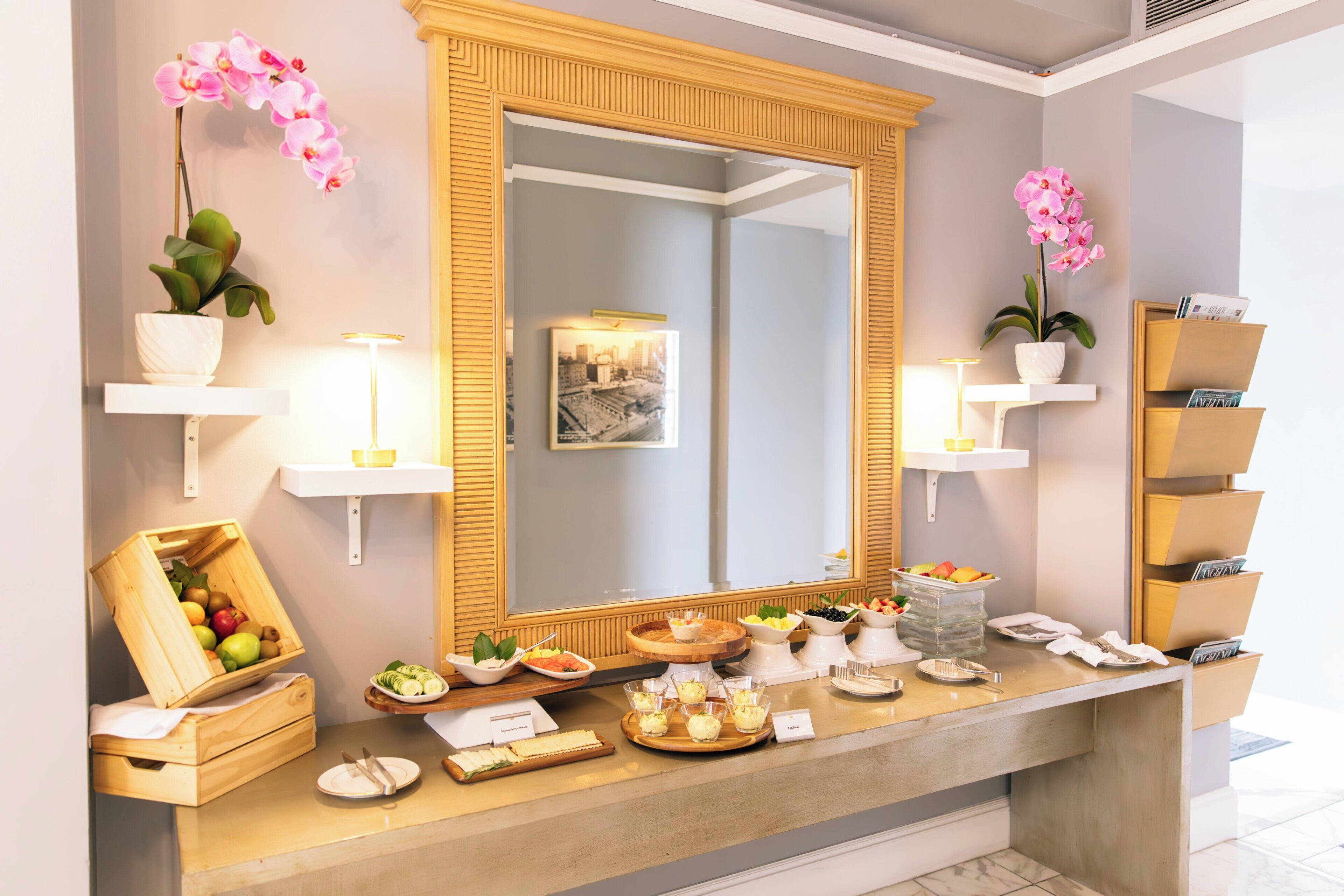 Zona de recepción de un hotel con un espejo grande, dos orquídeas en macetas, una exposición de frutas y un área de buffet.