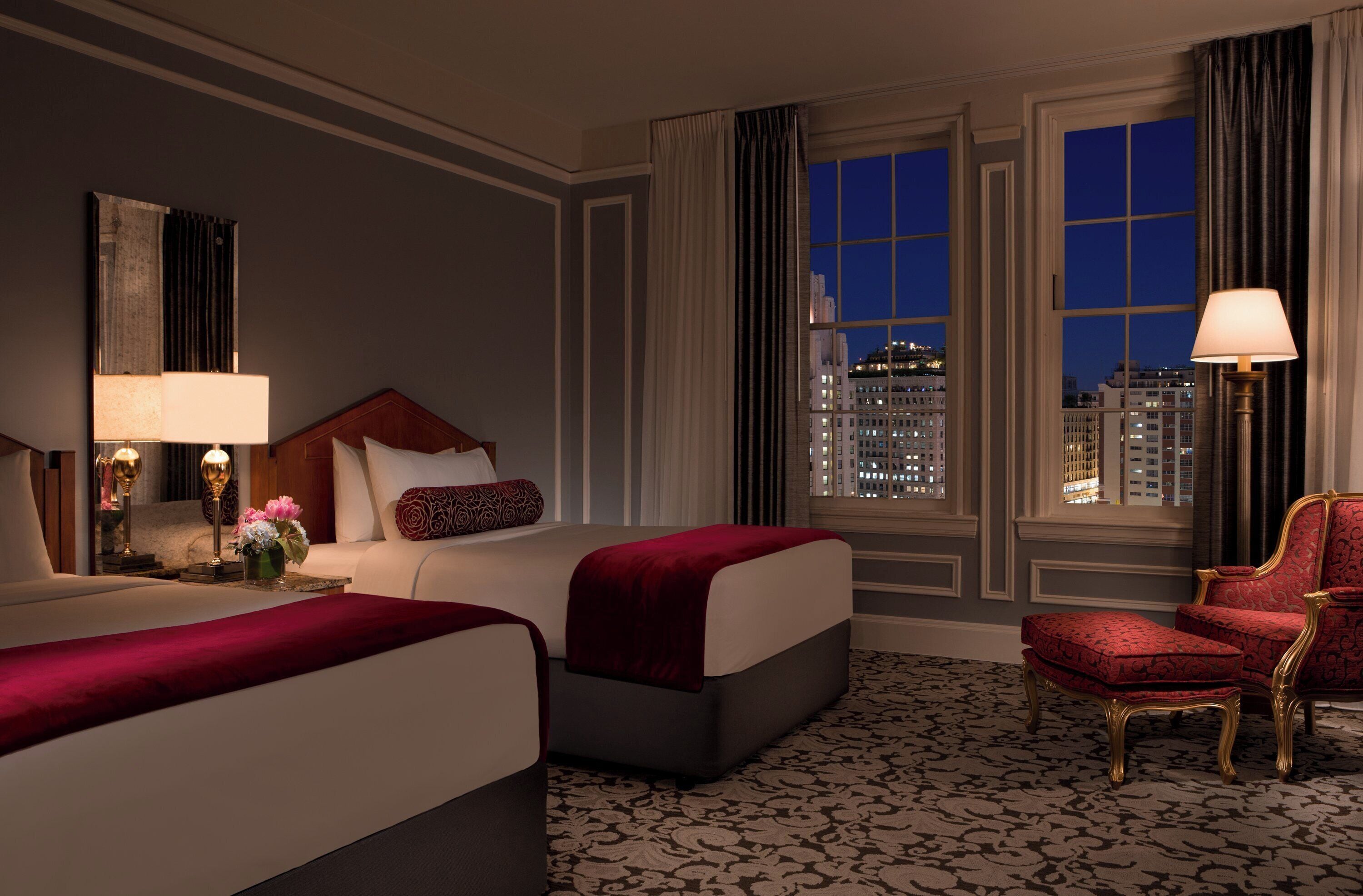 Habitación de hotel con dos camas, un área de descanso con una silla, una lámpara y vistas panorámicas de la ciudad a través de amplias ventanas.